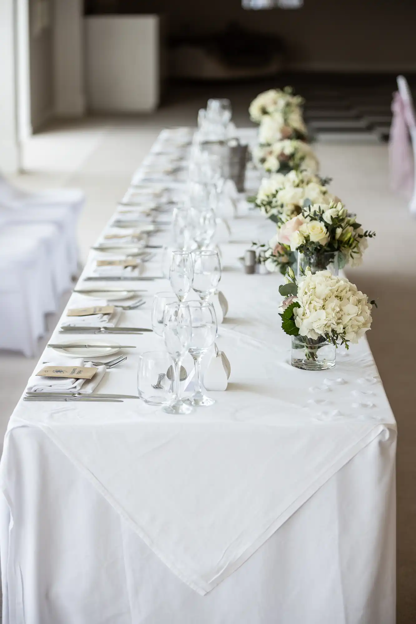 A long dining table is elegantly set with white linens, glassware, and floral centerpieces featuring white and cream flowers. The chairs are covered with white fabric.