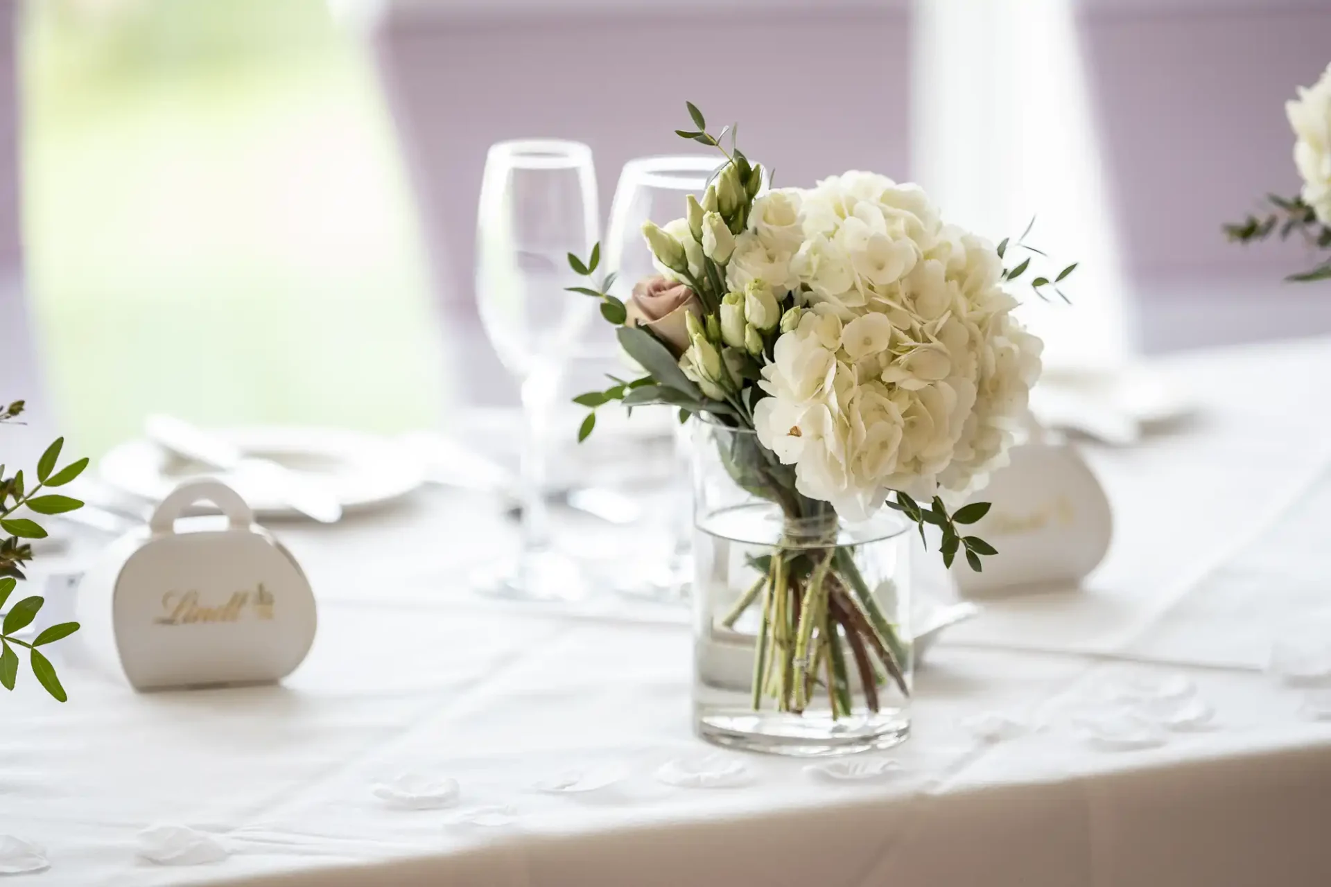 A clear vase with white flowers is on a table set for a meal, including wine glasses and white tableware.