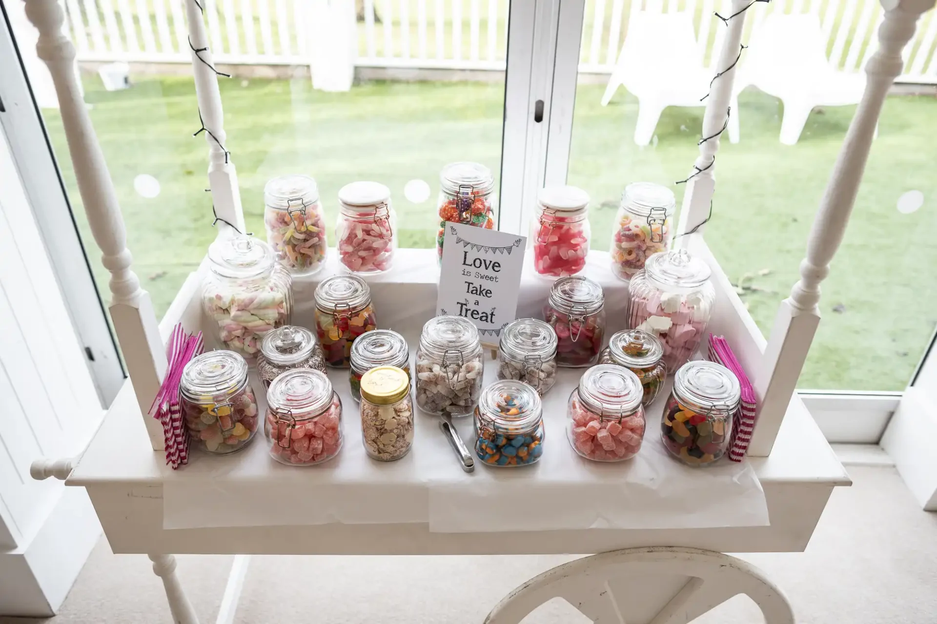 A candy cart with various jars filled with colorful candies, a small sign reading "Love is Sweet, Take a Treat," and pink striped bags for selection. It's positioned indoors near a window.