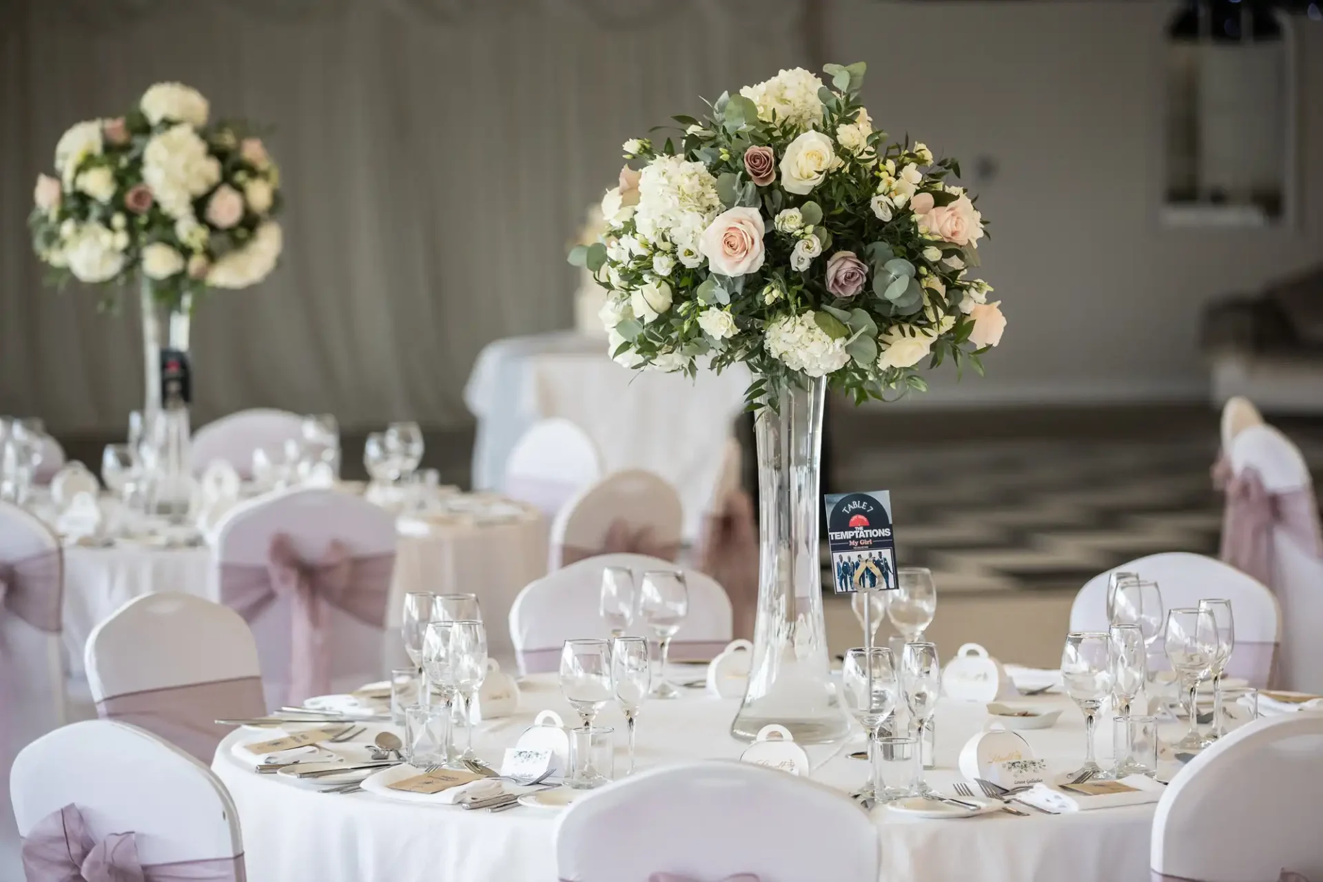 Elegant wedding reception setup with round tables, white tablecloths, tall floral centerpieces, and chairs adorned with pink sashes. Tables are set with glassware, napkins, and place cards.