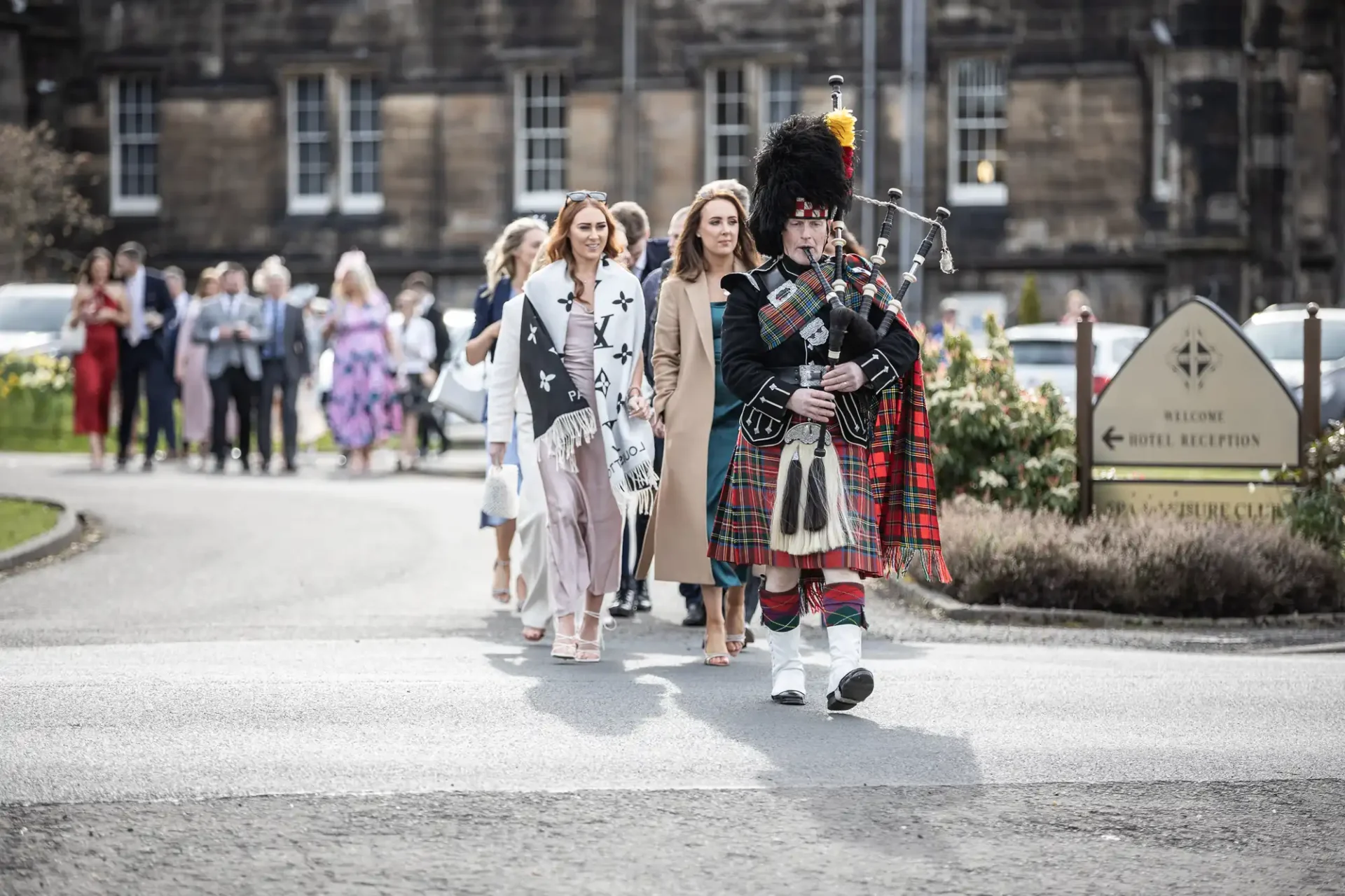 A bagpiper in traditional attire leads a procession of people down a street near a hotel reception sign.