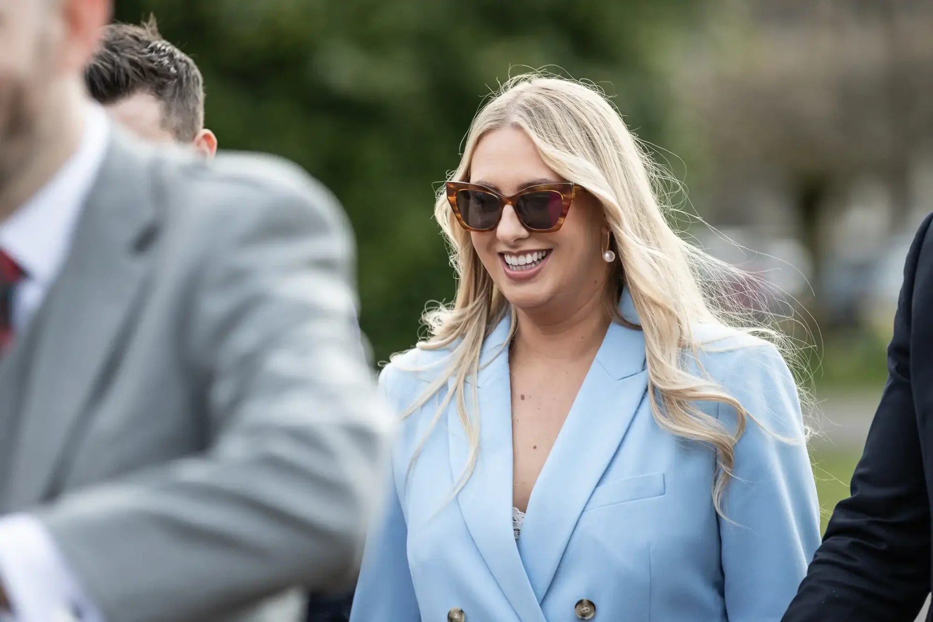 Smiling woman in sunglasses and a light blue jacket walks with a group outdoors.