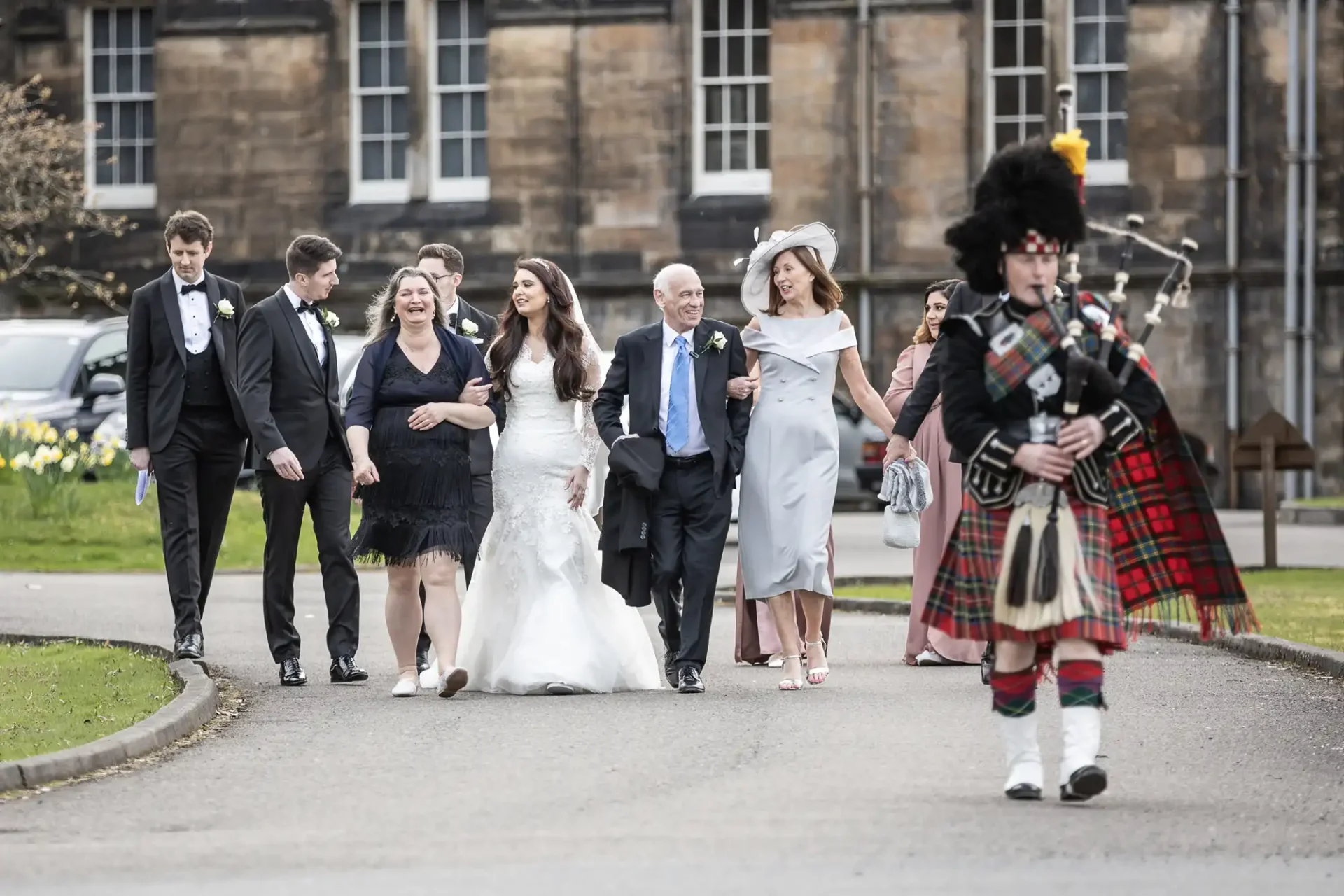 A bride and groom walk with a group of people, followed by a bagpiper in traditional attire, on a paved path in front of a historic building.