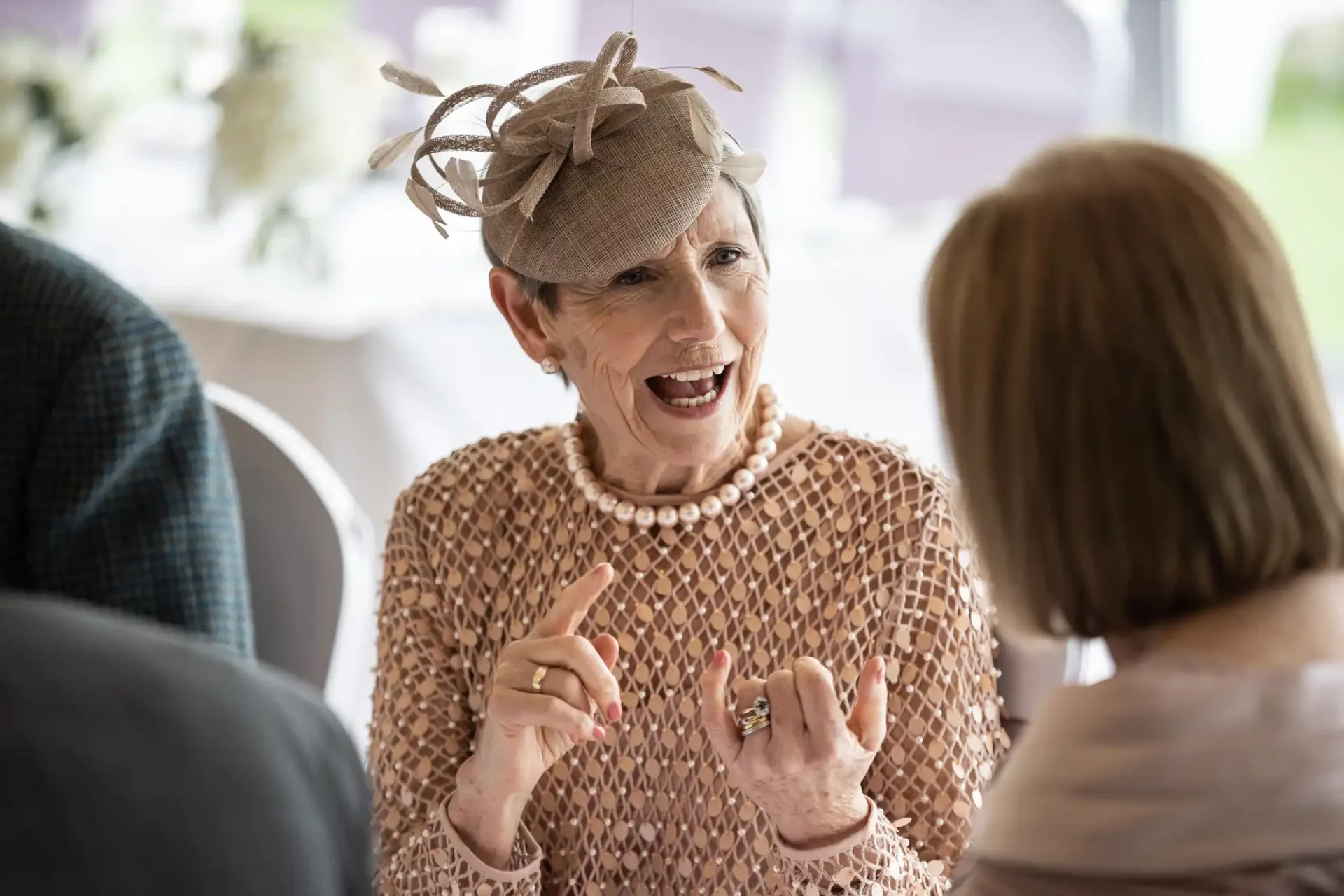 Elderly woman in textured dress and fascinator talking animatedly to another person indoors.