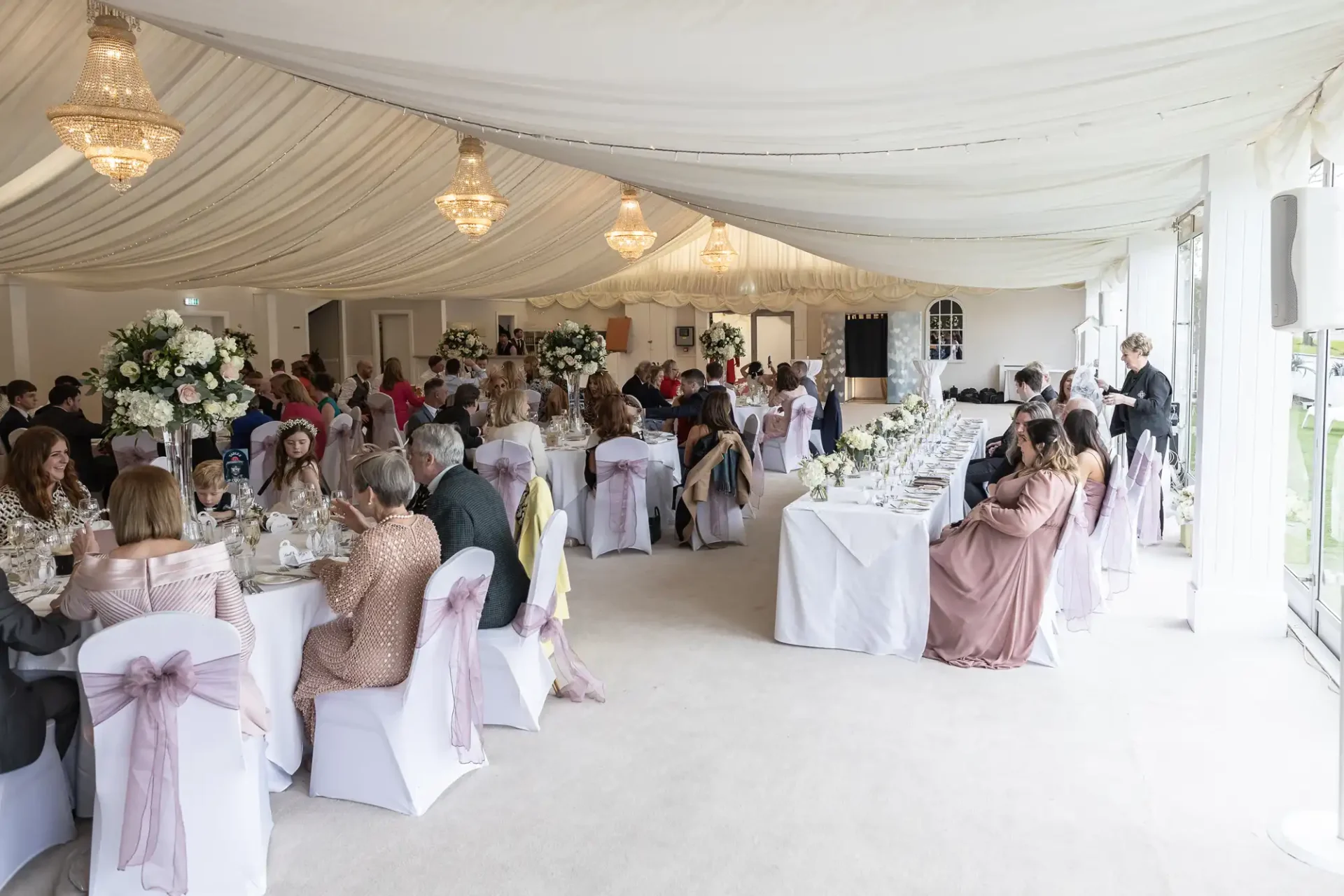 Guests seated at tables inside a decorated event tent with white drapes and chandeliers, attending what appears to be a formal gathering or celebration.