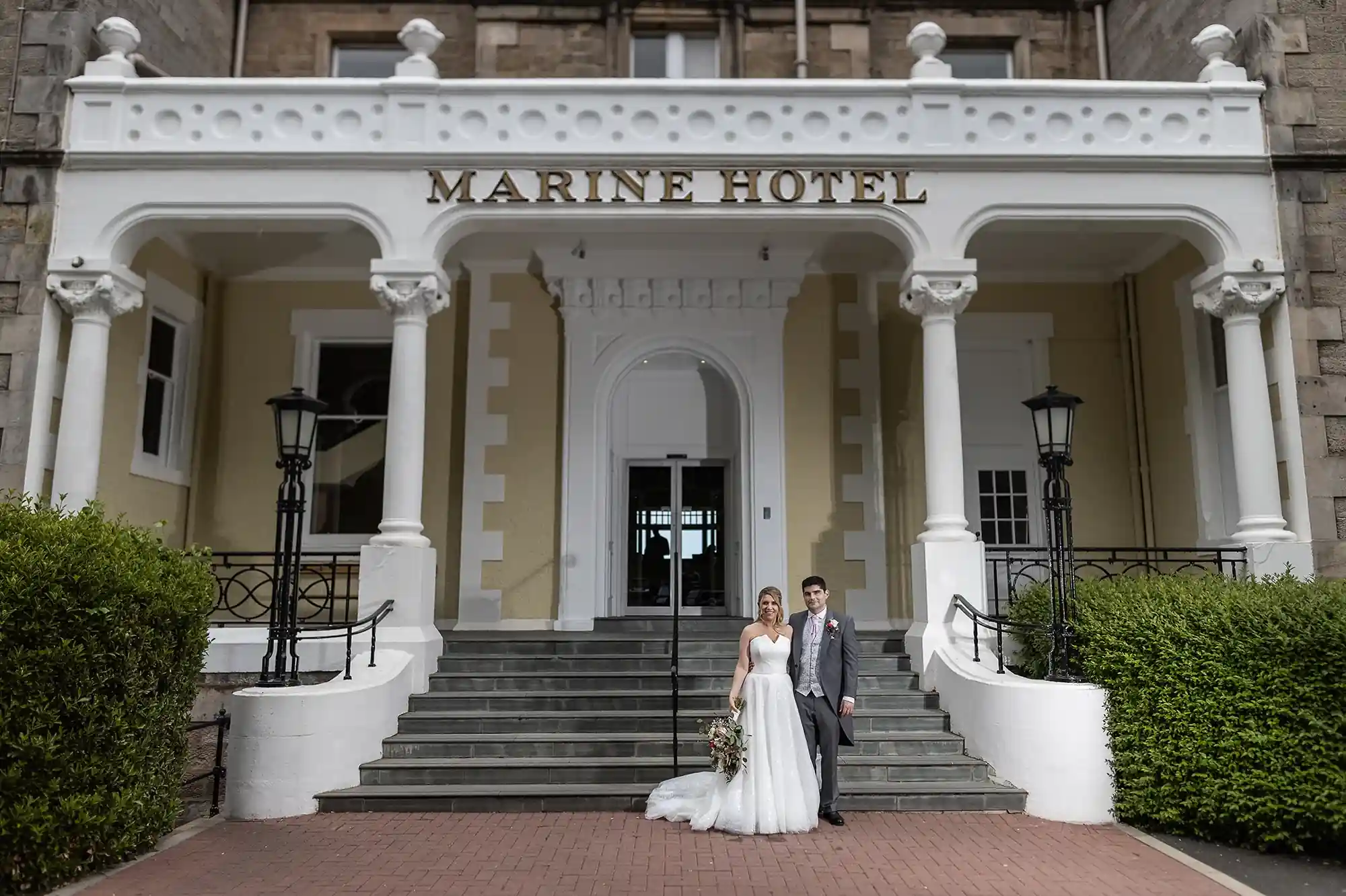 A newlywed couple standing on steps outside the marine hotel, with the groom in a suit and the bride in a white gown holding a bouquet.