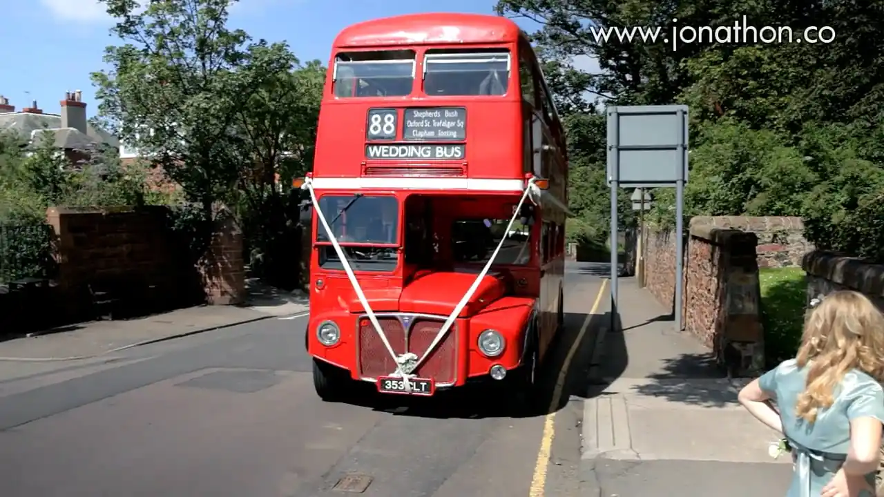 Marine Hotel Wedding Videographer - Bridal Preparations image of a red double-decker bus decorated with white ribbons and a "Wedding Bus" sign is parked at the side of a street on a sunny day.