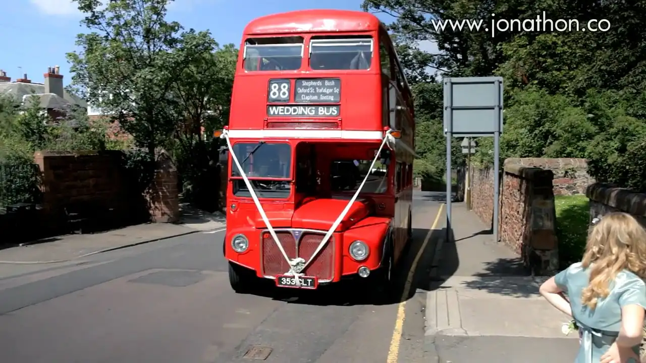 A red double-decker bus decorated with white ribbons and a "Wedding Bus" sign is parked on a quiet street next to stone walls and trees.