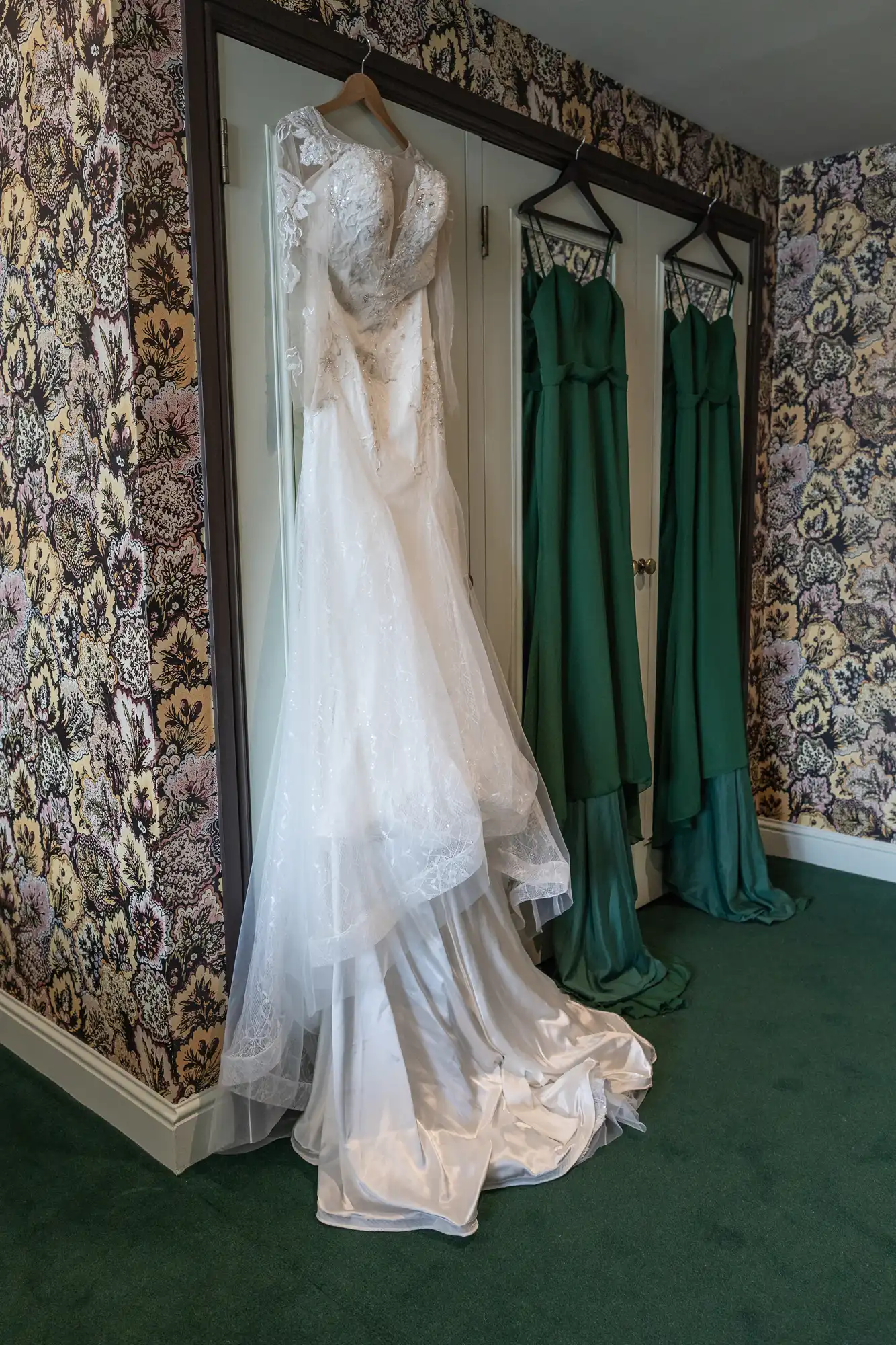 A white wedding dress with lace details hangs beside two dark green bridesmaid dresses in a room with floral wallpaper.