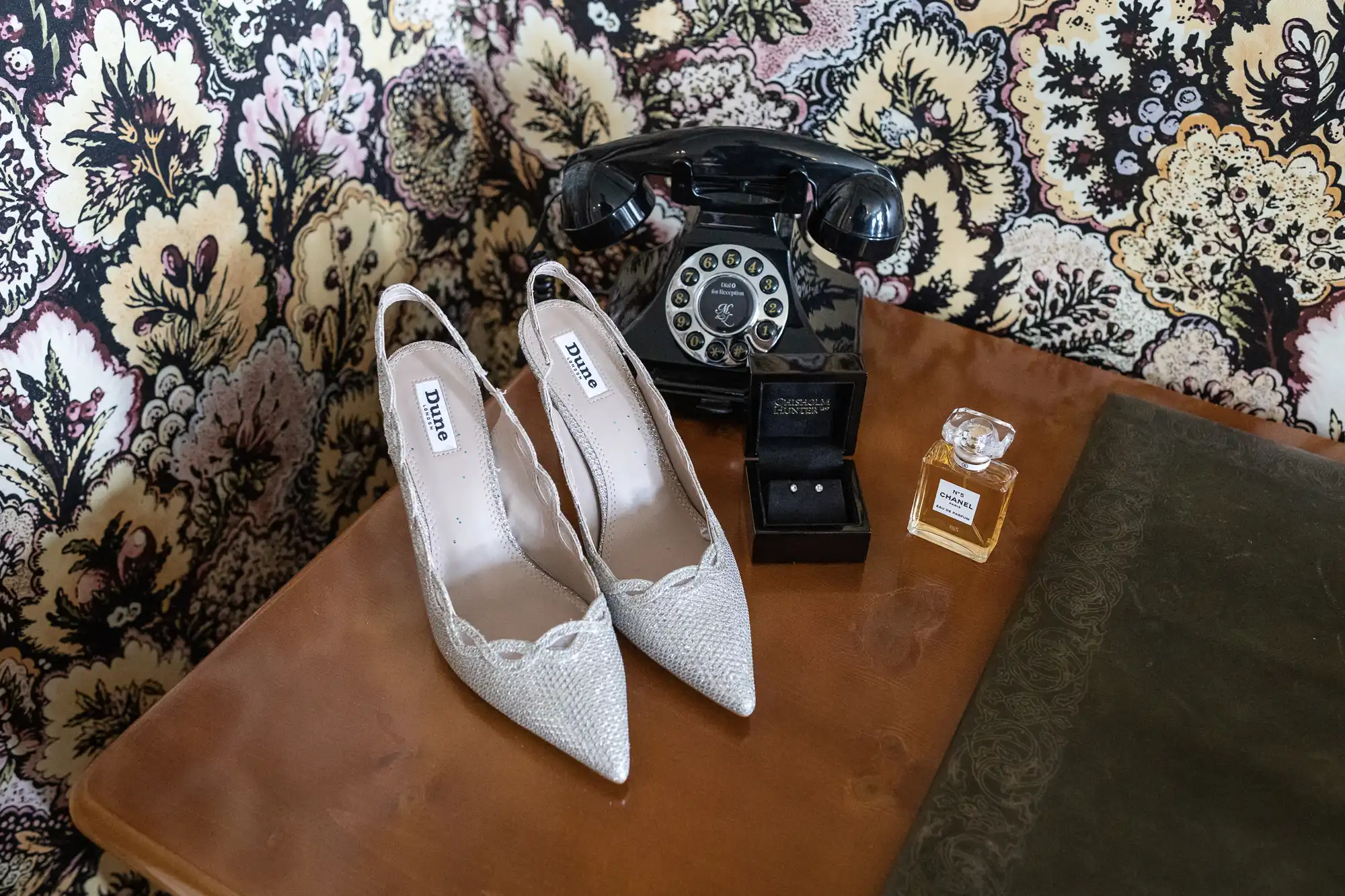 A pair of white bridal shoes, a vintage black telephone, a watch, and a perfume bottle on a brown leather surface against a floral backdrop.