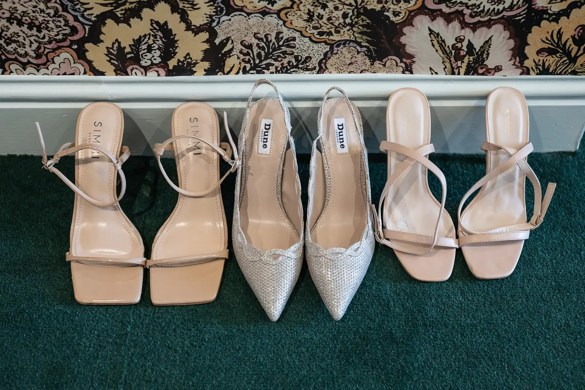 Three pairs of women's shoes, including beige heels and silvery pointed heels, neatly aligned on a green carpet against a floral wall.
