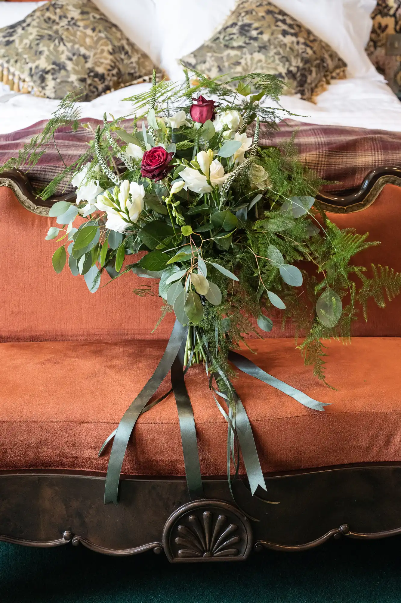 A bouquet of red roses and white flowers with green foliage and long ribbons, placed on a plush orange bedspread with patterned pillows in the background.