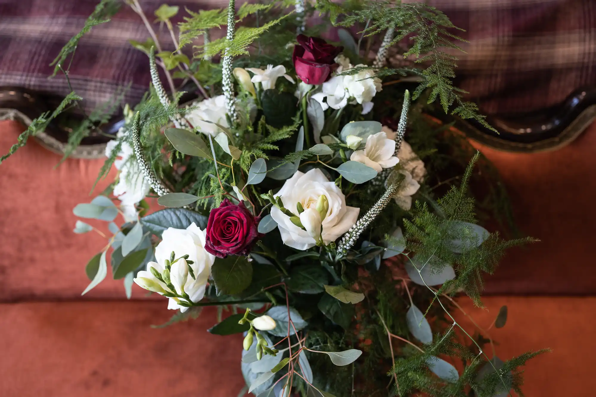 A close-up of a floral arrangement featuring white roses, dark red roses, and various greenery on a plaid fabric background.