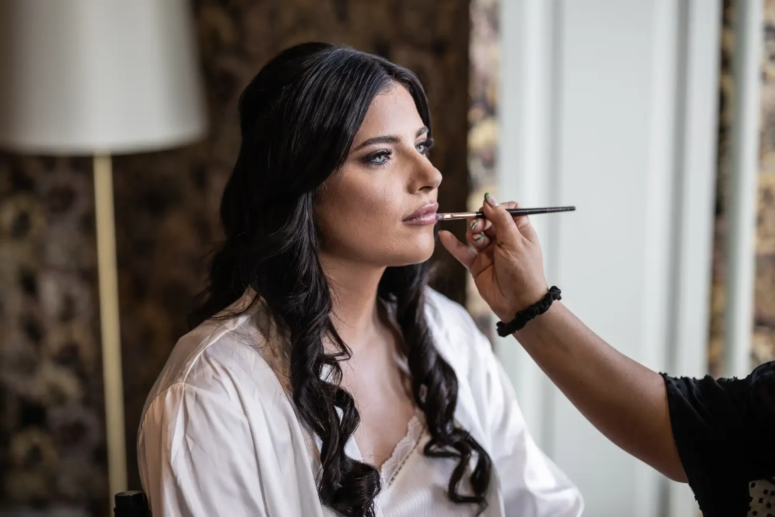 A woman in a white robe getting her makeup done with a brush by another person in a dimly lit room.