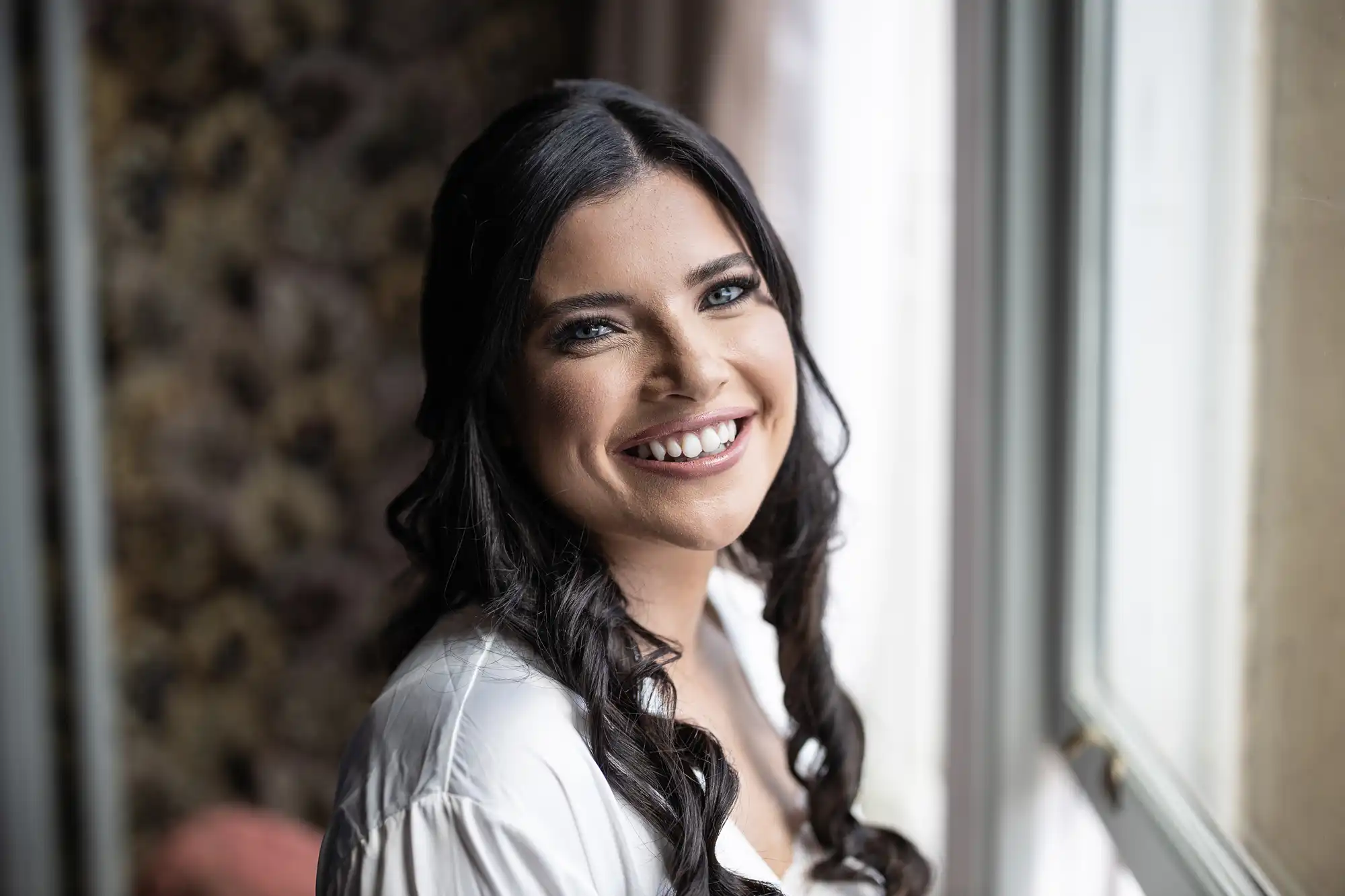 A smiling woman with long braided hair, wearing a white blouse, looks joyfully towards the camera by a window.