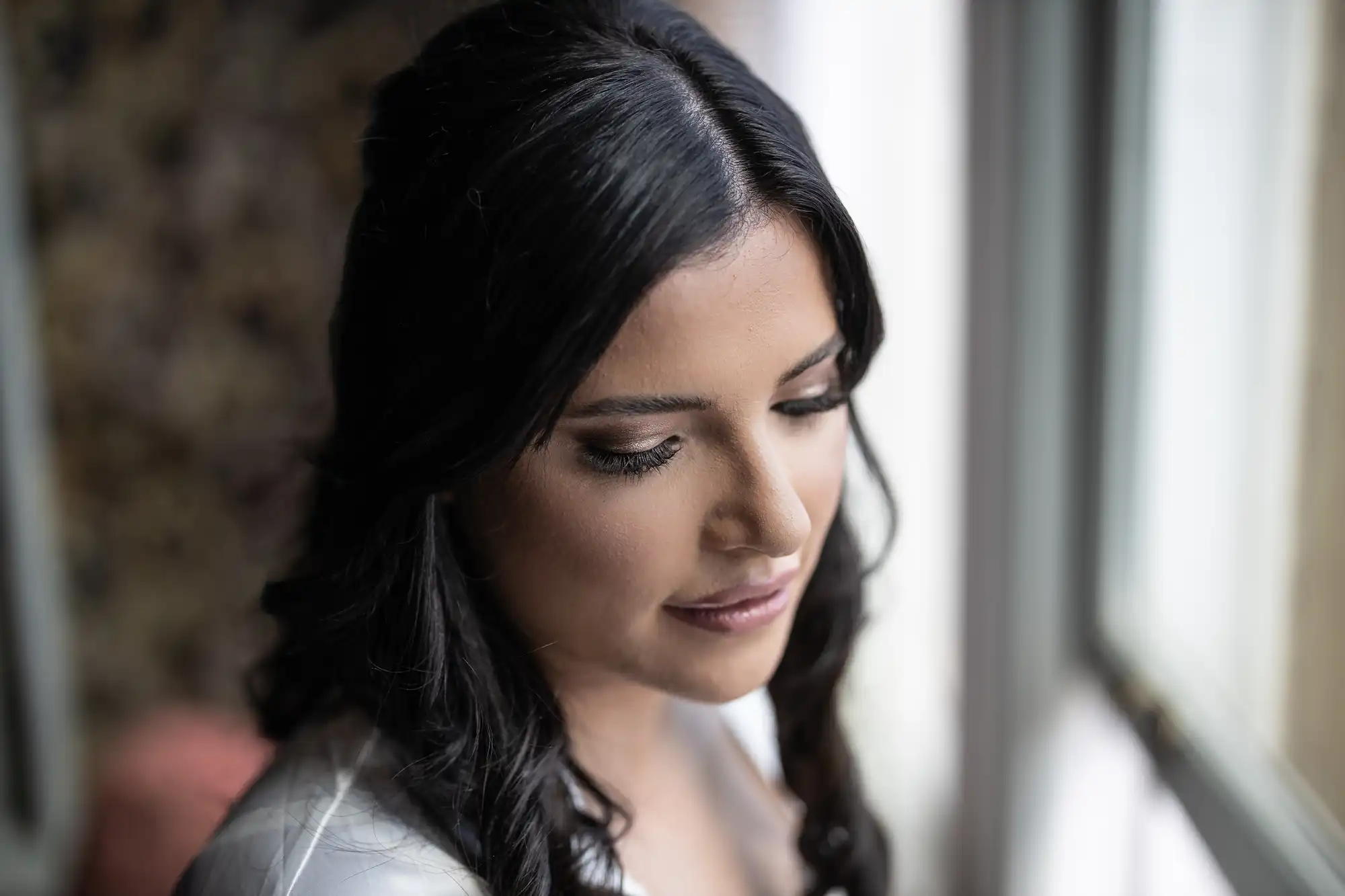 Woman with dark hair in white attire gazing down thoughtfully near a window.