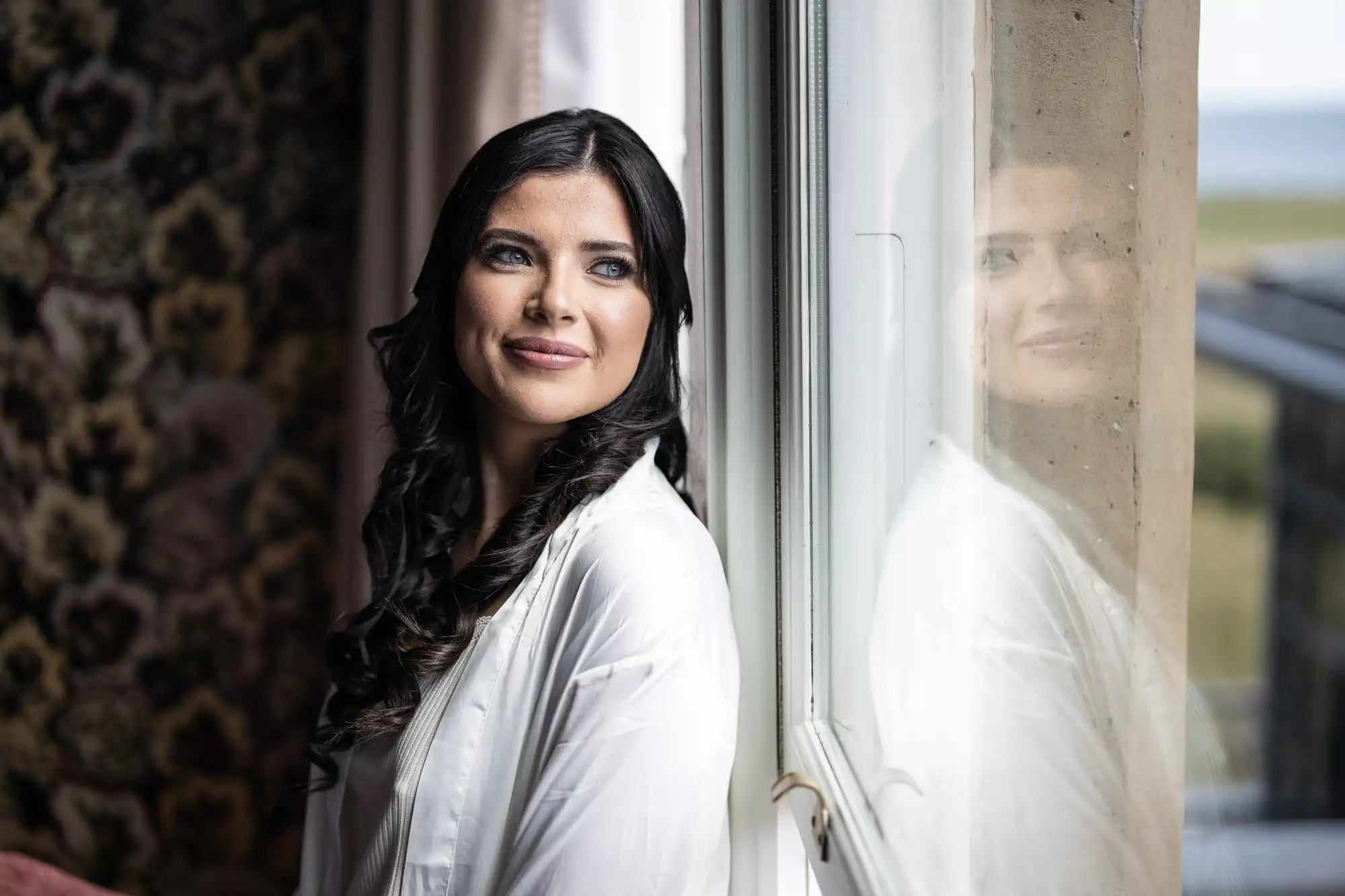 A woman with dark hair, wearing a white robe, smiling and looking away from the camera, with her reflection visible in a window beside her.