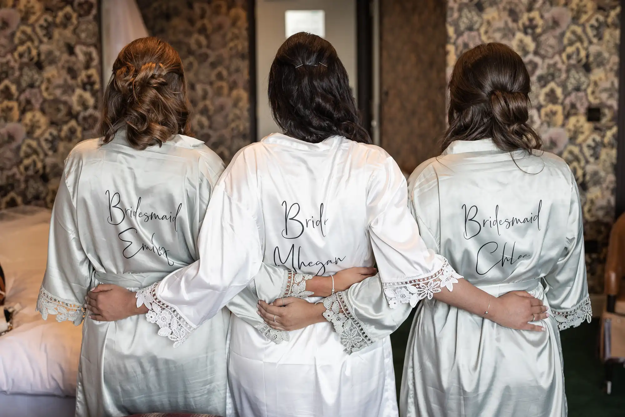 Three women wearing silk robes with "Bridesmaid" and "Bride" written on the back, standing together with arms around each other, facing away from the camera.