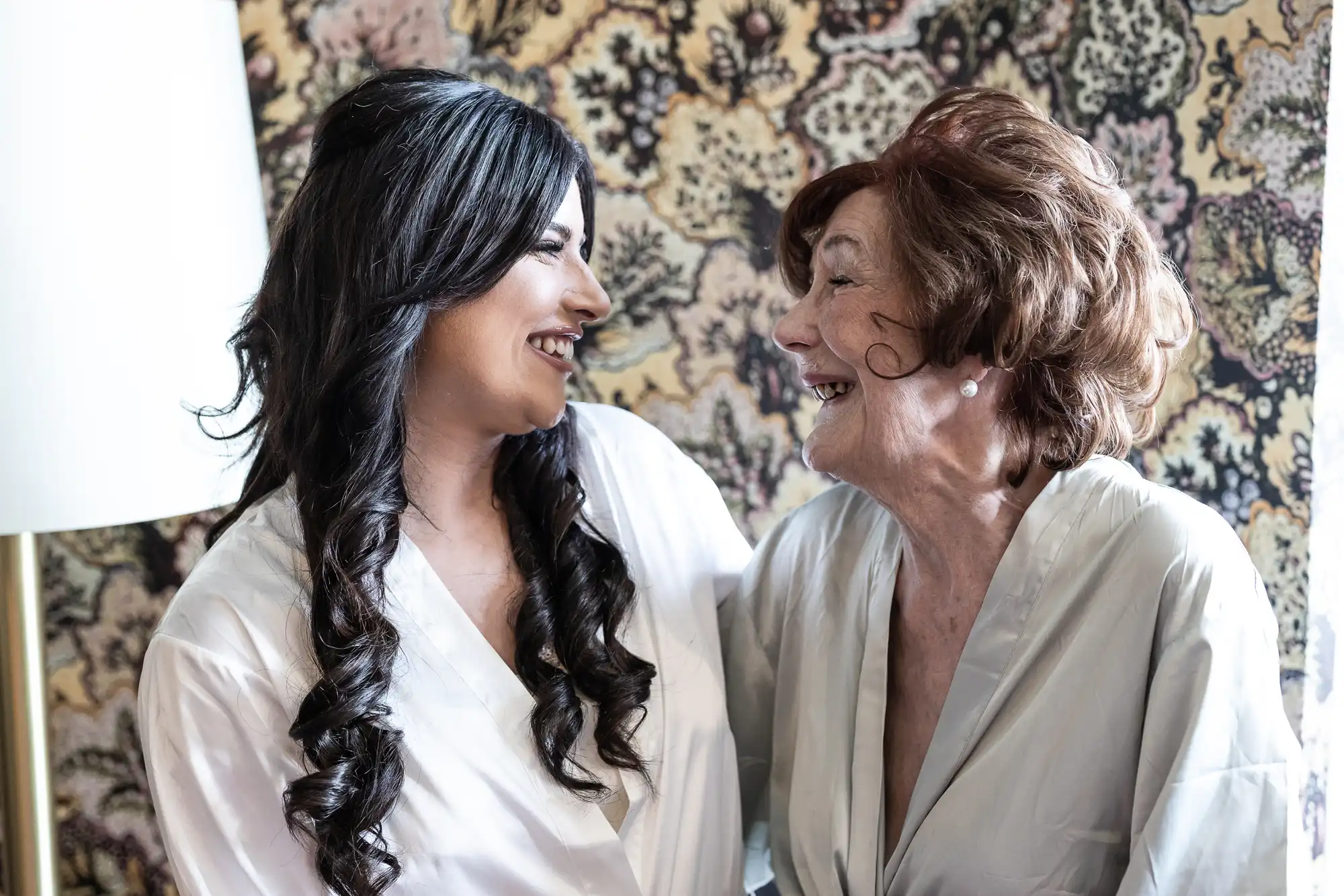 Two women, one younger and one older, smiling at each other in a warmly lit room with patterned wallpaper, both wearing white robes.