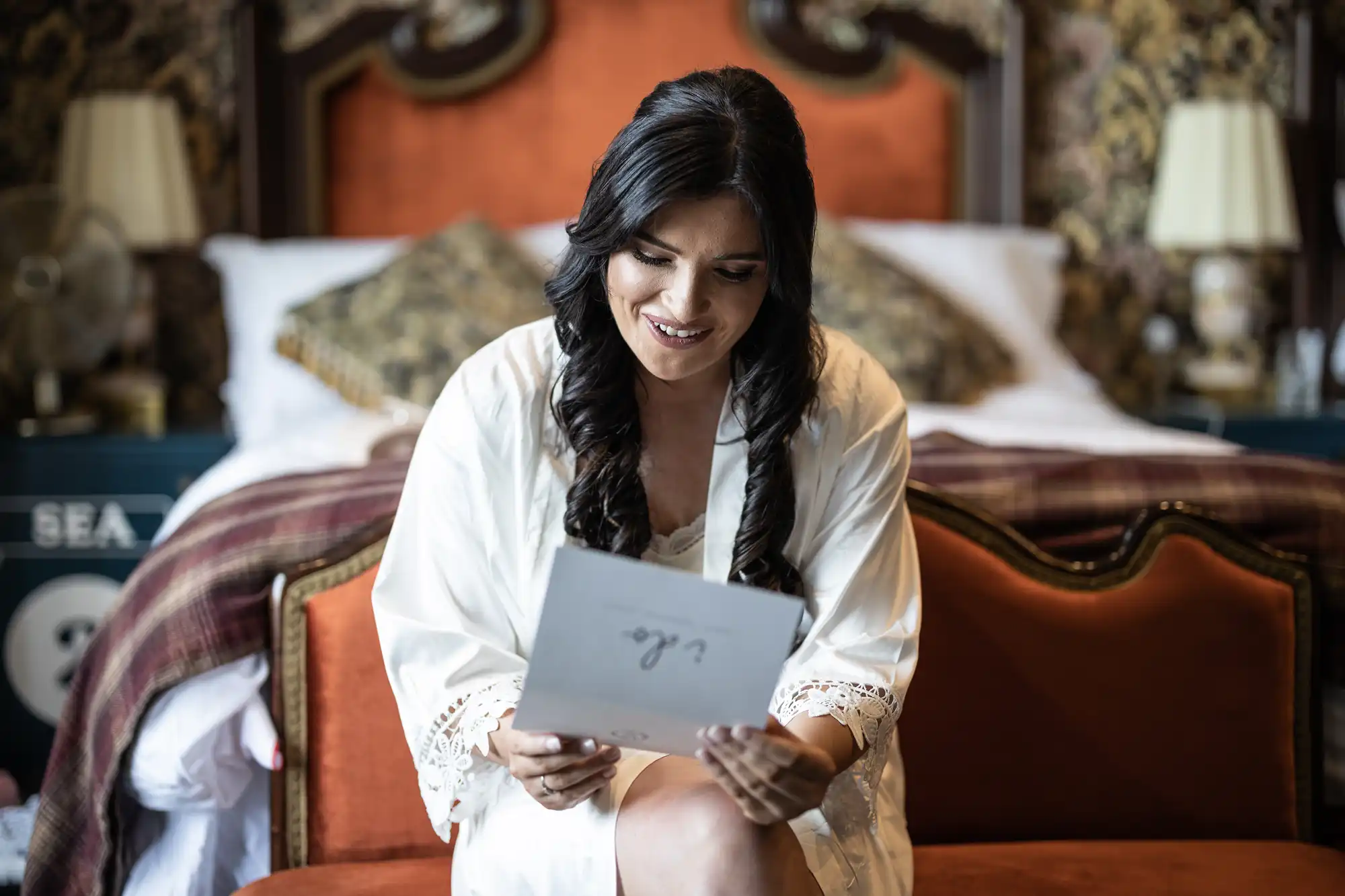 A woman in a white robe sitting on a red couch in an ornately decorated room, smiling as she reads a letter.