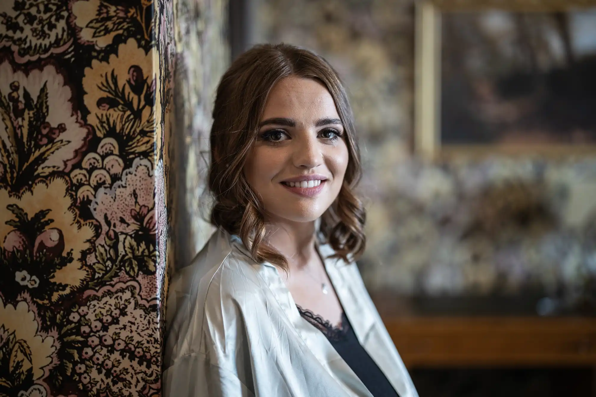 A smiling woman with shoulder-length brown hair, wearing a white blouse, seated next to a floral-patterned wall.