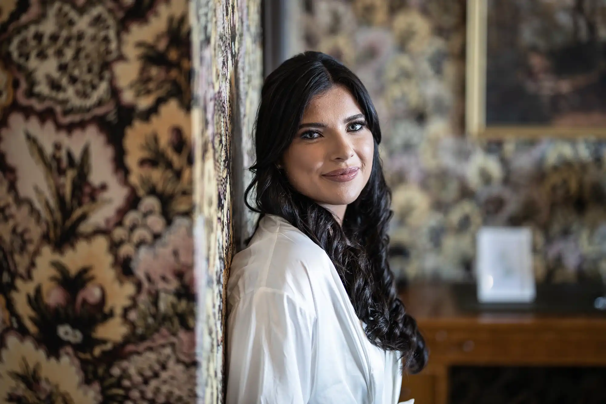 A woman with dark hair and a white blouse smiles gently, leaning against a wall with ornate tapestry in an elegantly decorated room.