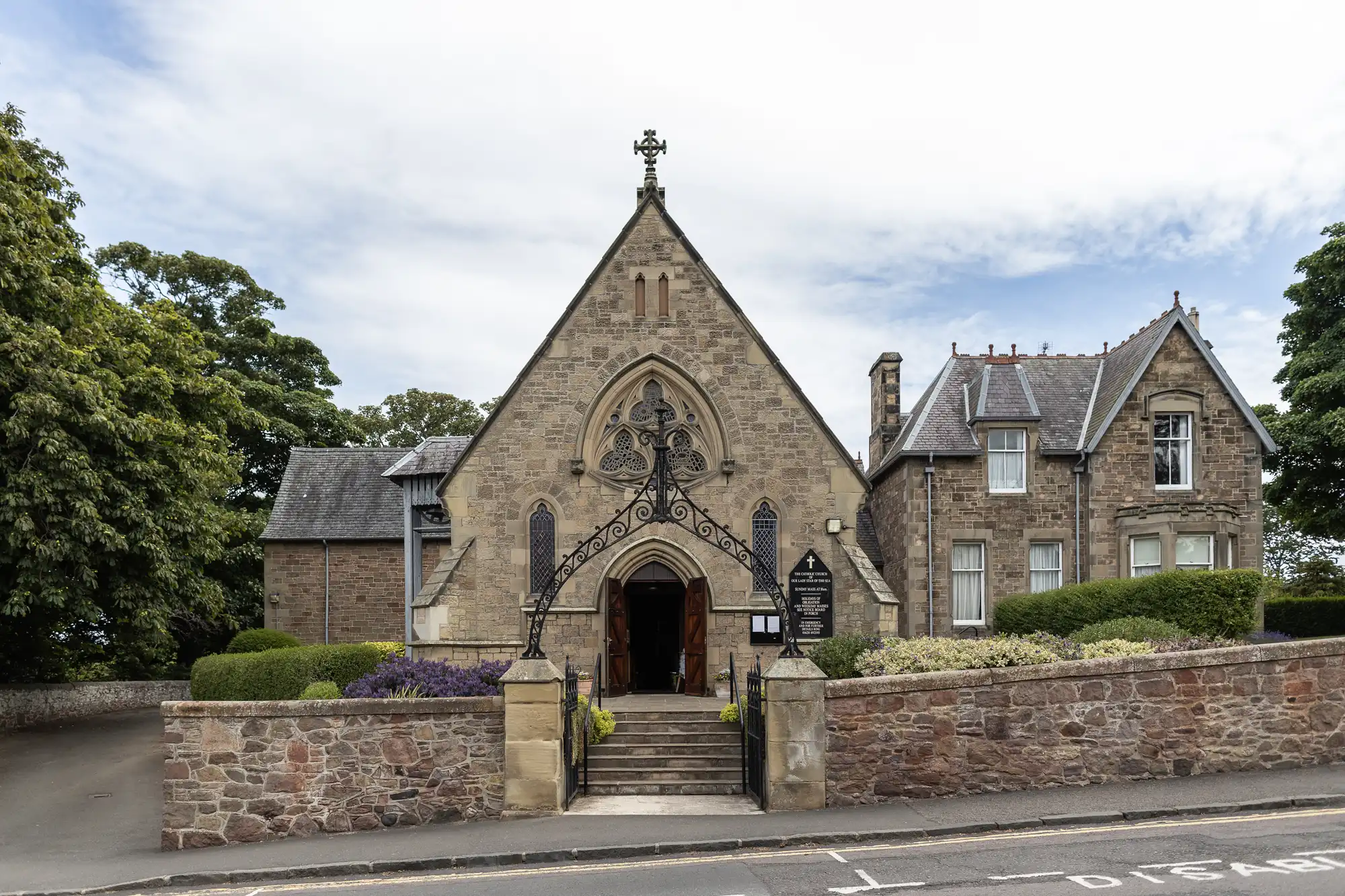 A quaint stone church with an arched entrance and detailed façade, flanked by a traditional house, set against a clear sky and surrounded by lush greenery.