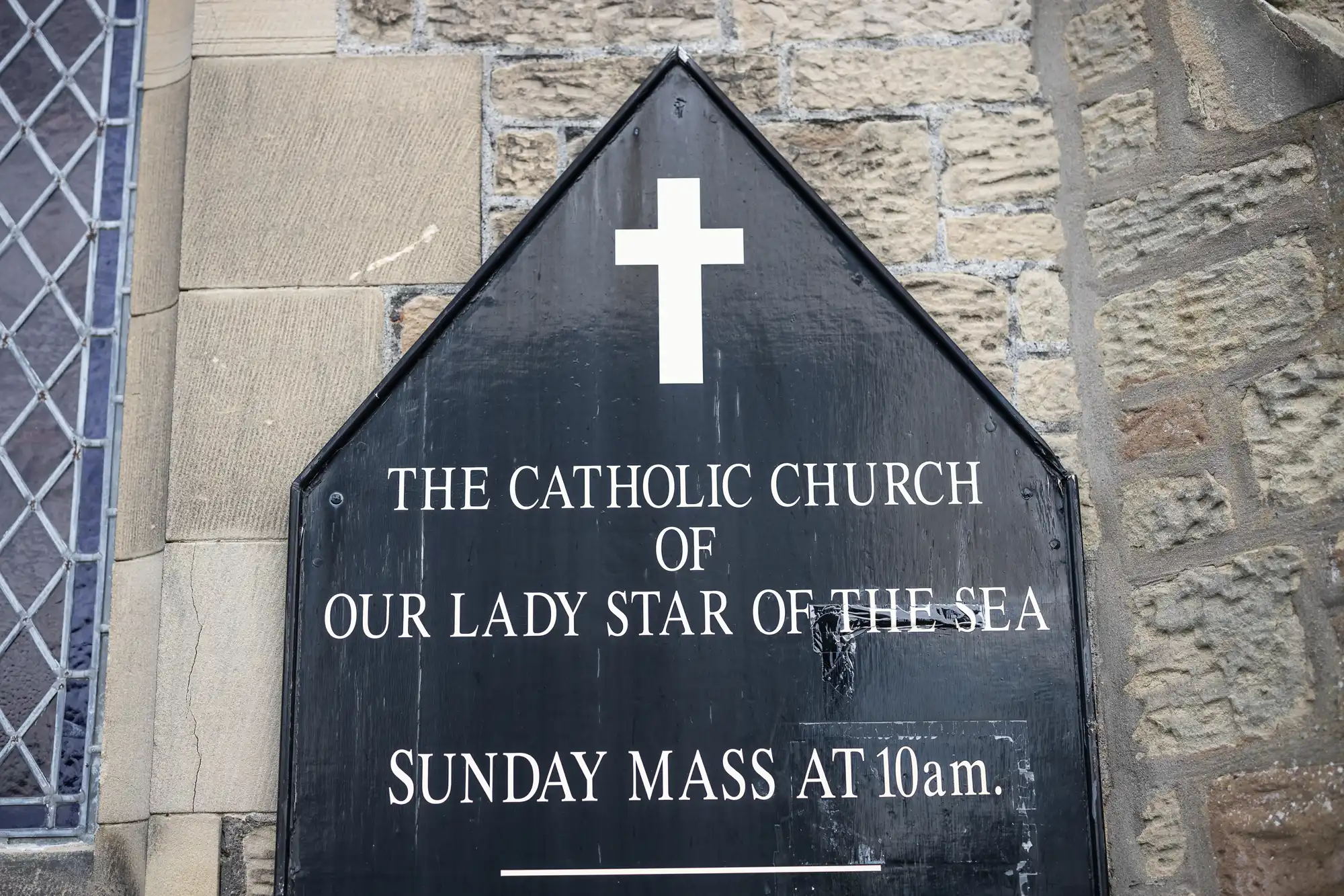 Black triangular sign with white text "The Catholic Church of Our Lady Star of the Sea - Sunday Mass at 10am." with a white cross above the text, mounted on a stone wall.