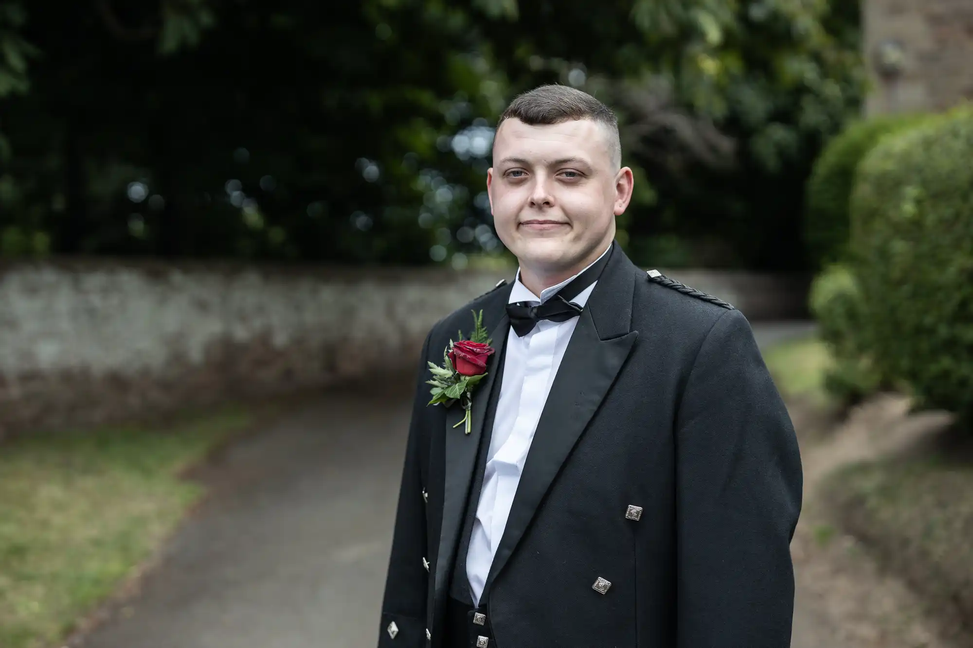 A young man in a tuxedo with a boutonniere smiles subtly, standing on a pathway with greenery in the background.
