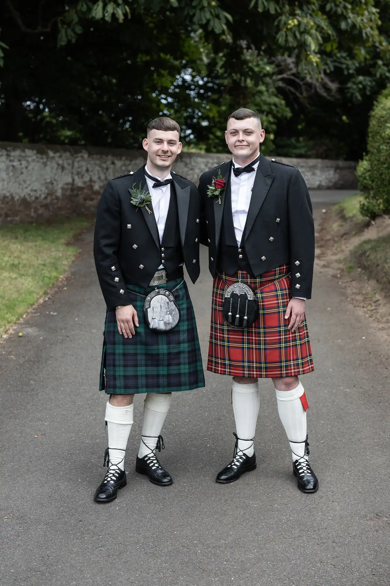 Two men in traditional Scottish attire, including kilts and sporran, smiling and standing on a path lined with greenery.
