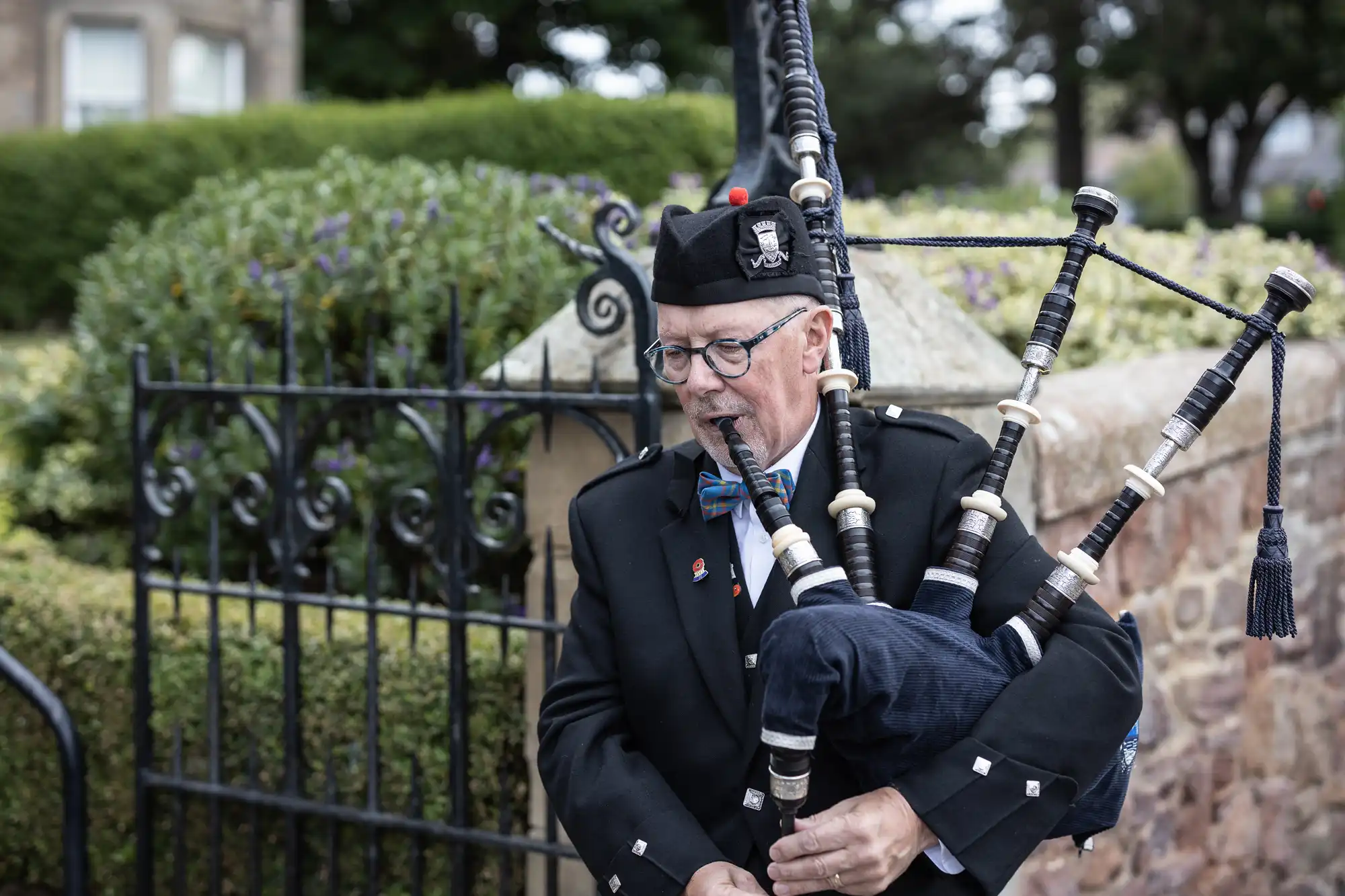 A man in traditional Scottish attire playing bagpipes outdoors near a metal fence and flowerbeds.