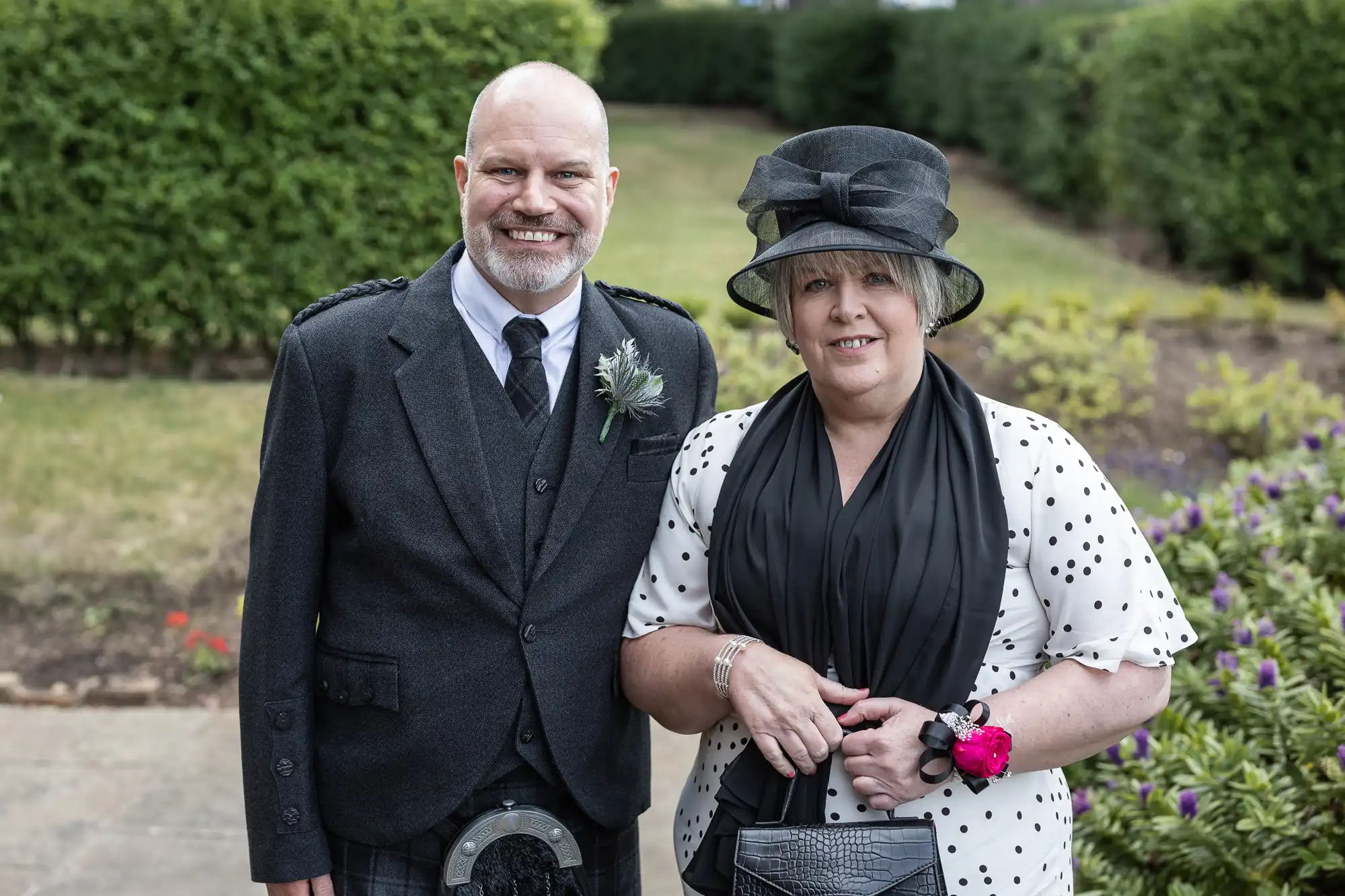 A man in a kilt and a woman in a black and white dress with hats, smiling in a garden at a formal event.