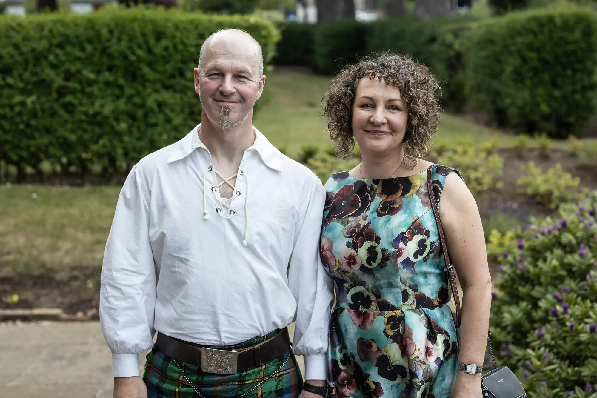 A man in a kilt and a woman in a floral dress standing together in a garden.