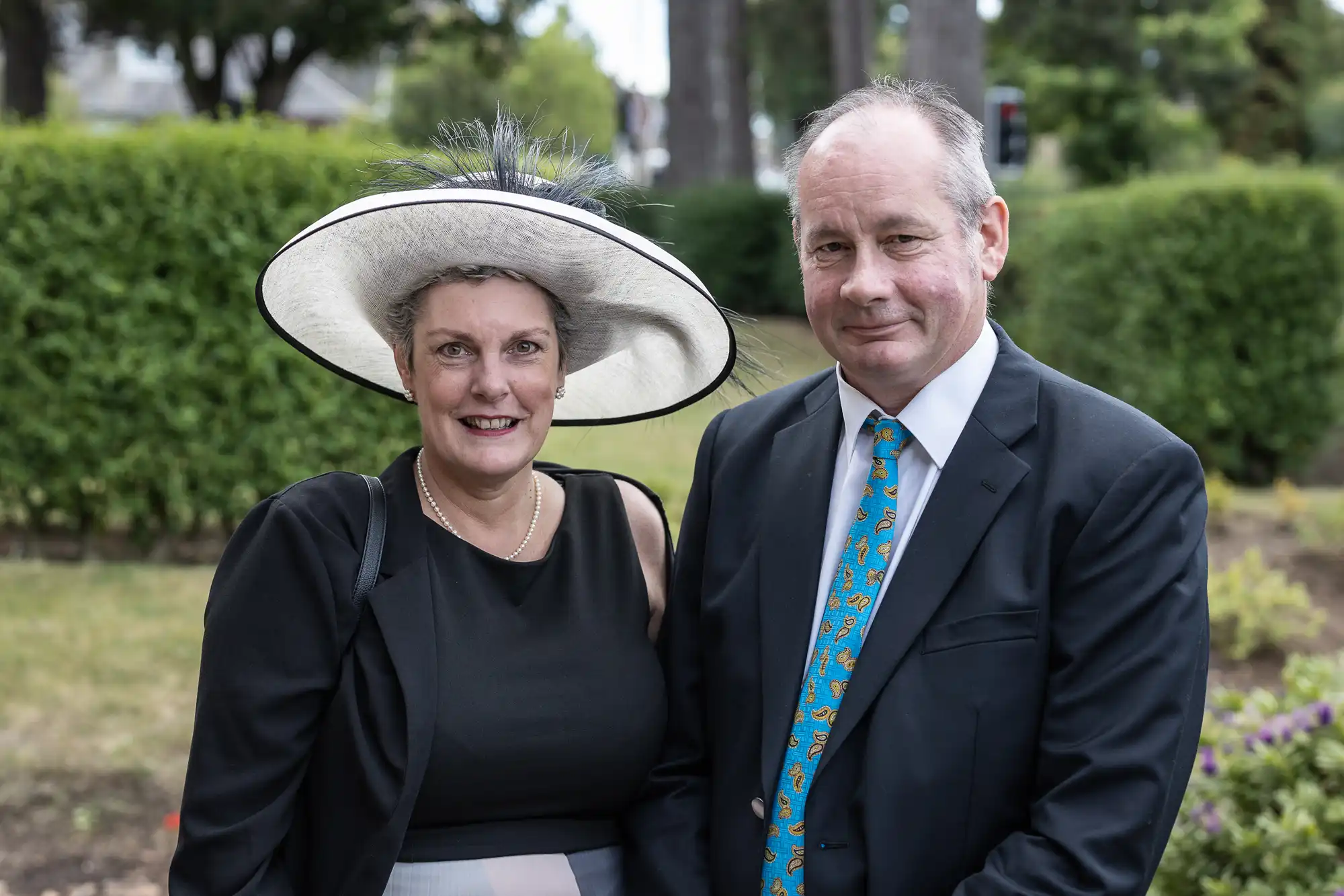 A mature couple, the woman wearing a wide-brimmed hat and black dress, the man in a navy suit with a patterned tie, standing together in a garden.