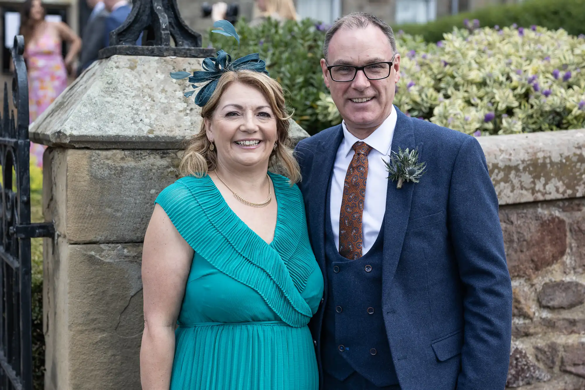 A smiling couple posing in formal attire at an outdoor event, the woman in a teal dress and fascinator, the man in a navy suit and patterned tie.