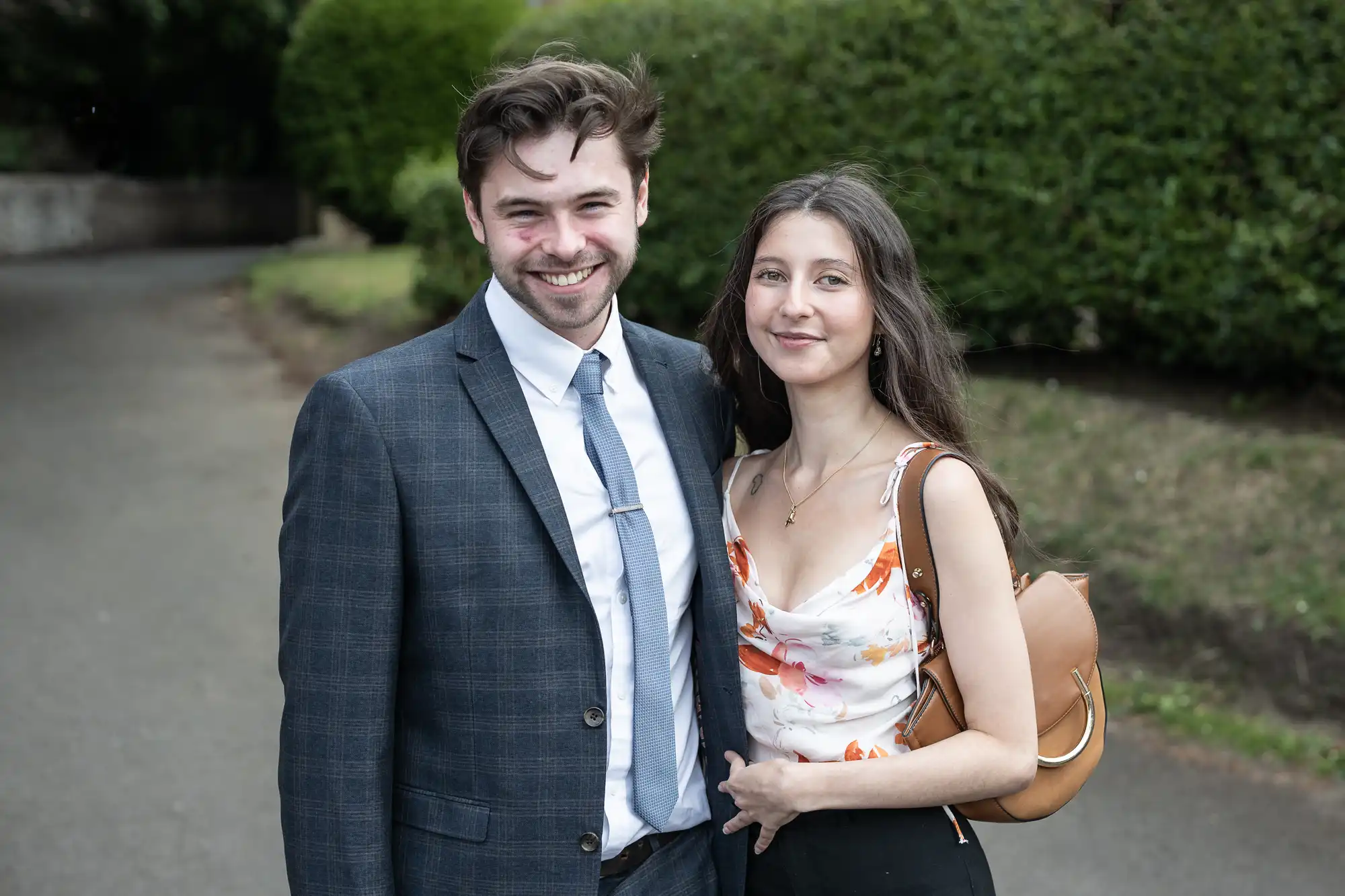 A smiling couple, the man in a grey suit and the woman in a floral dress, standing on a path with green bushes in the background.