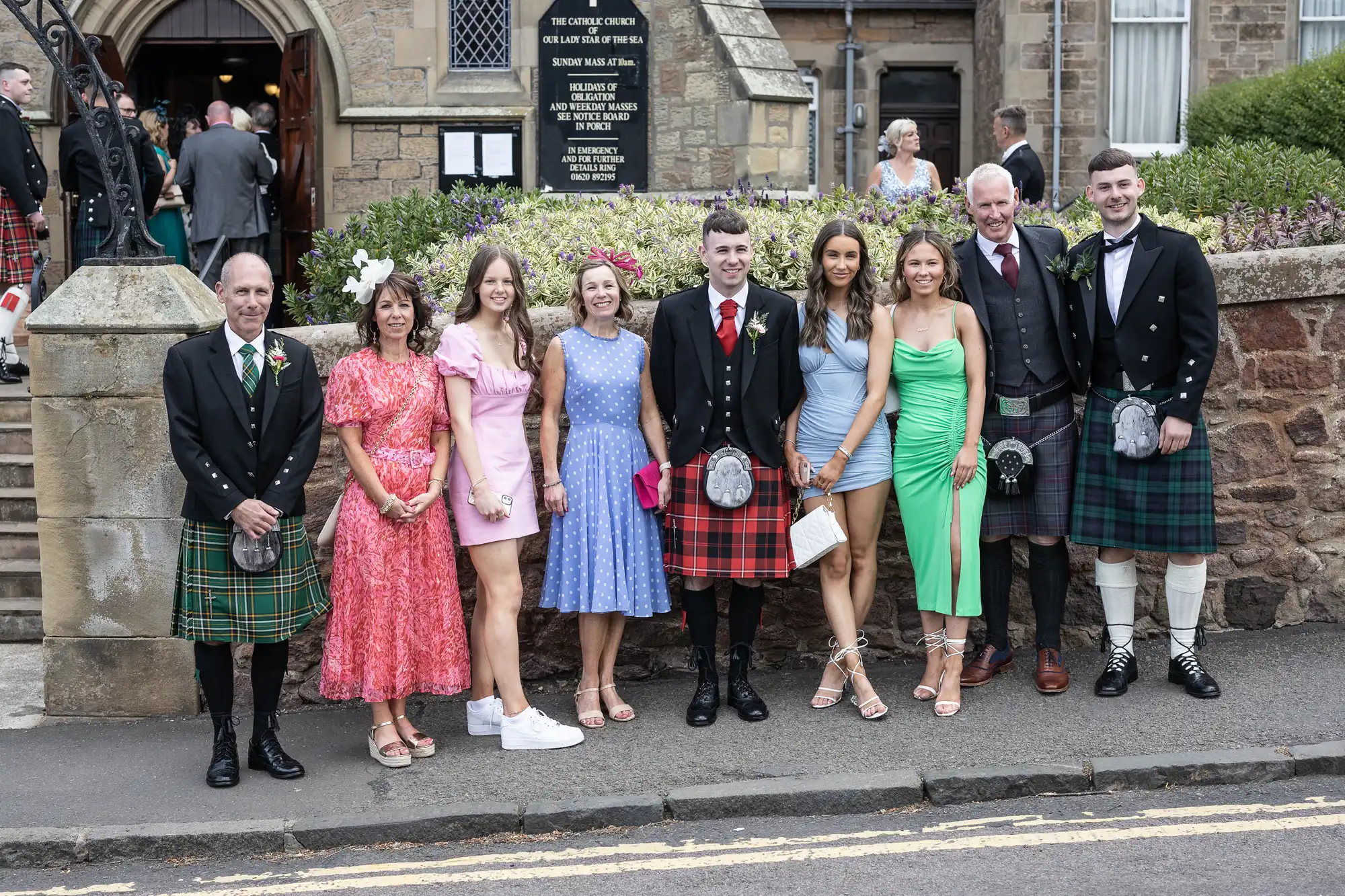 A group of nine people dressed semi-formally, with some men in kilts, posing outside a church entrance.