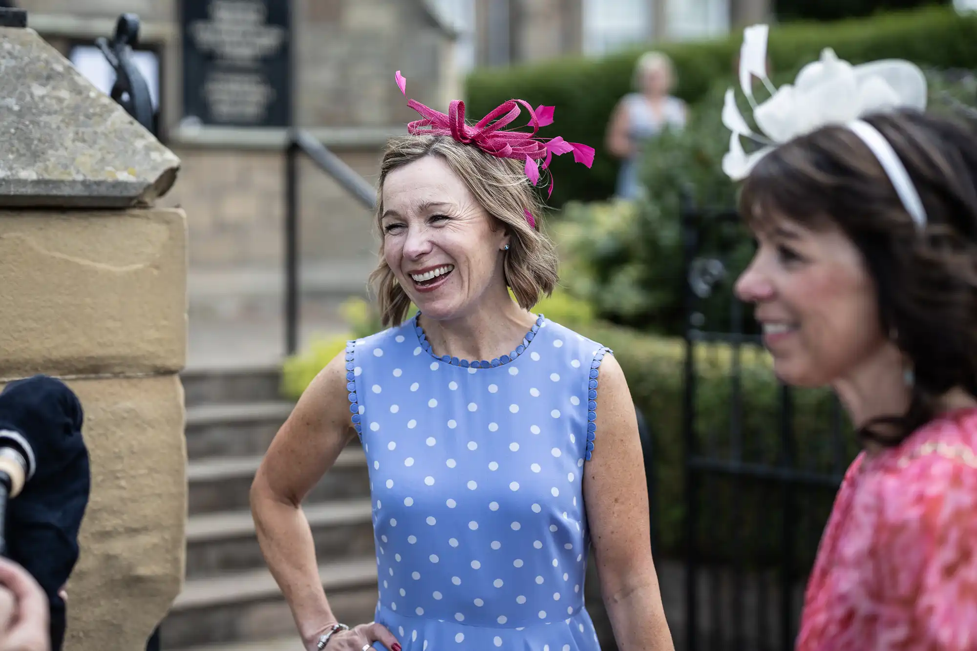 A woman in a blue polka dot dress and a pink fascinator laughs joyfully at a social gathering, accompanied by another woman in a white hat.
