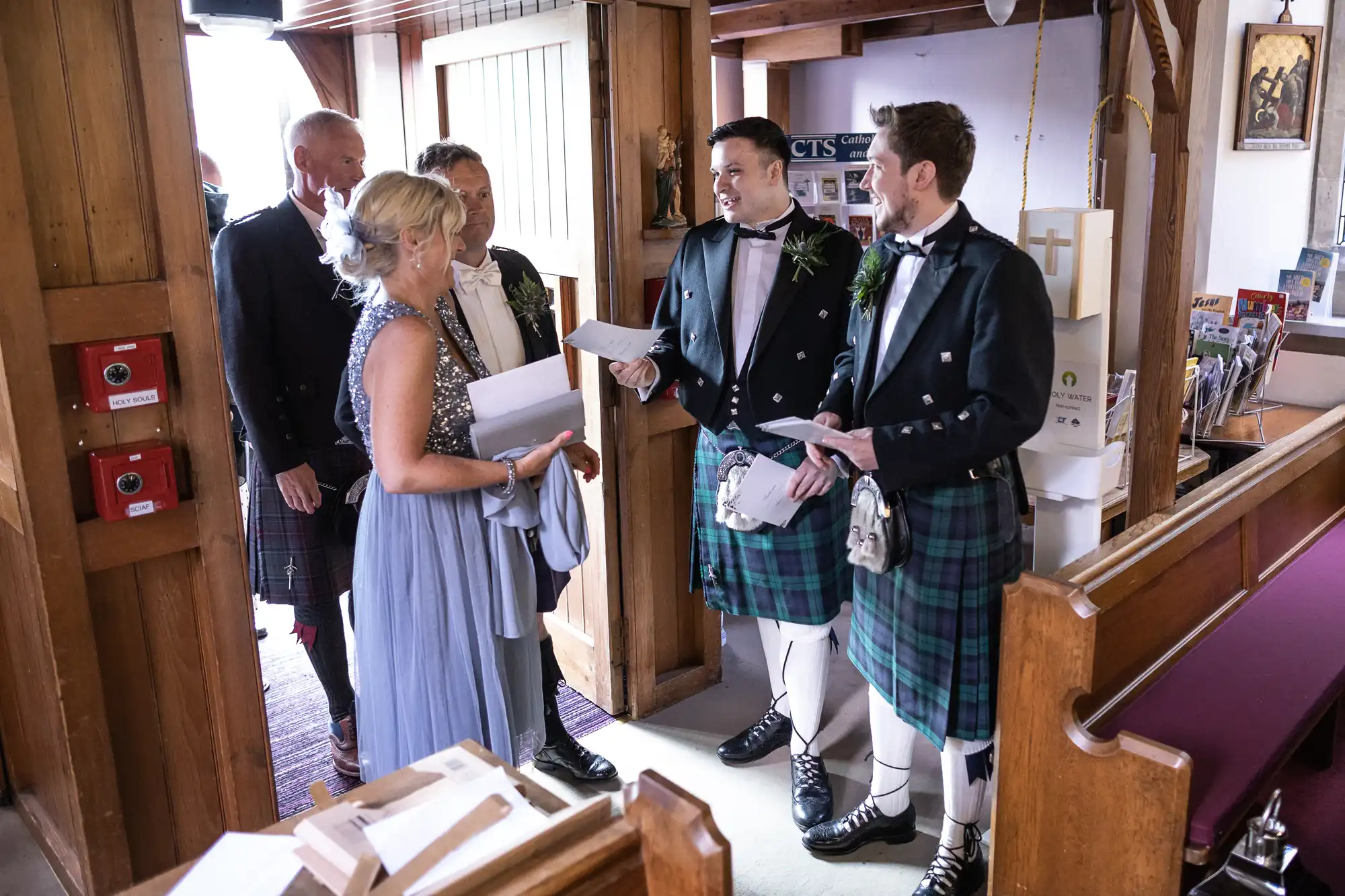 Two couples exchanging vows in a church, with one couple wearing traditional kilts. Guests stand nearby inside a wooden interior.