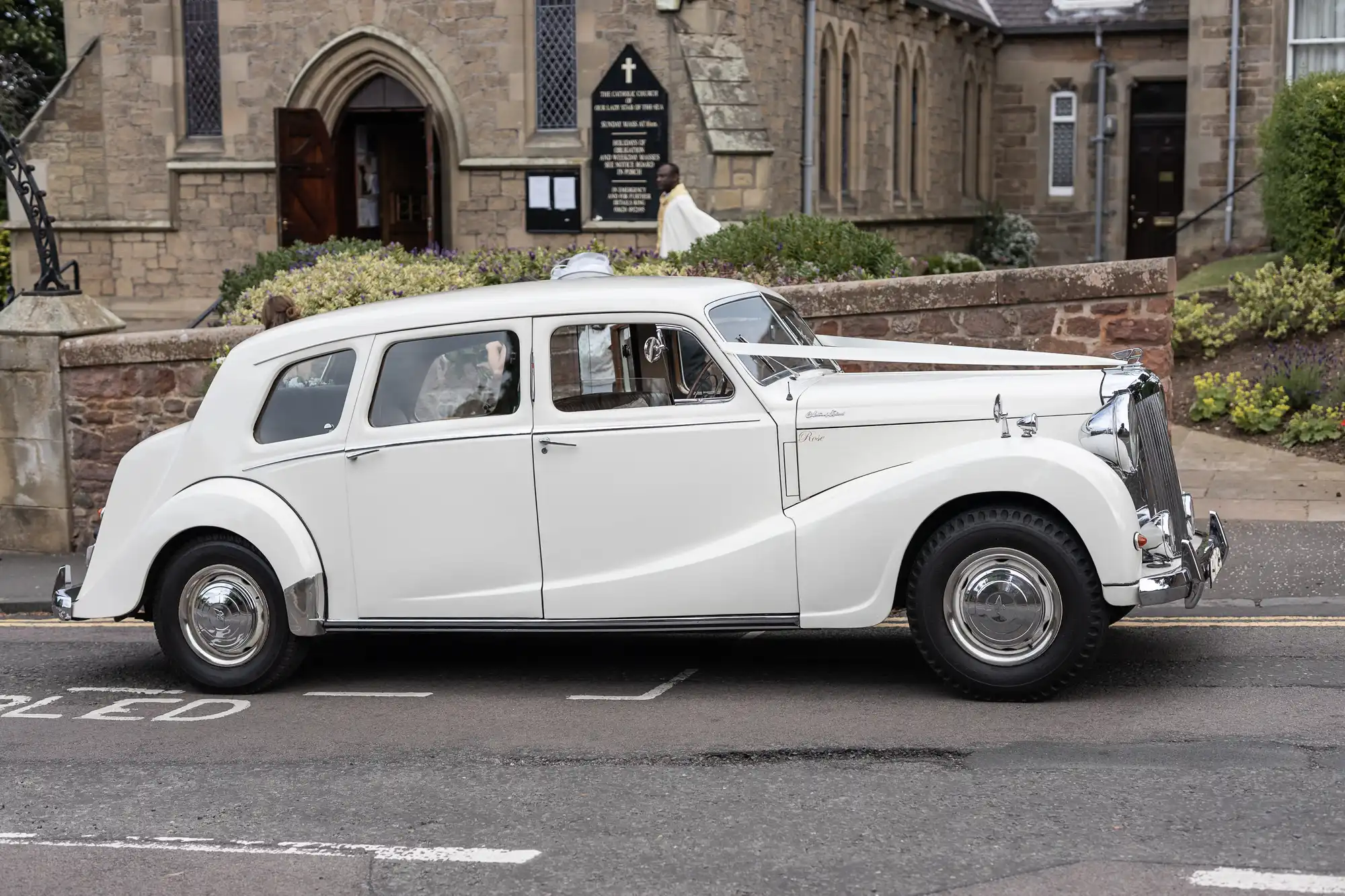 A vintage white Rolls-Royce parked on a street, with a bride visible in the rear seat, looking out of the window.