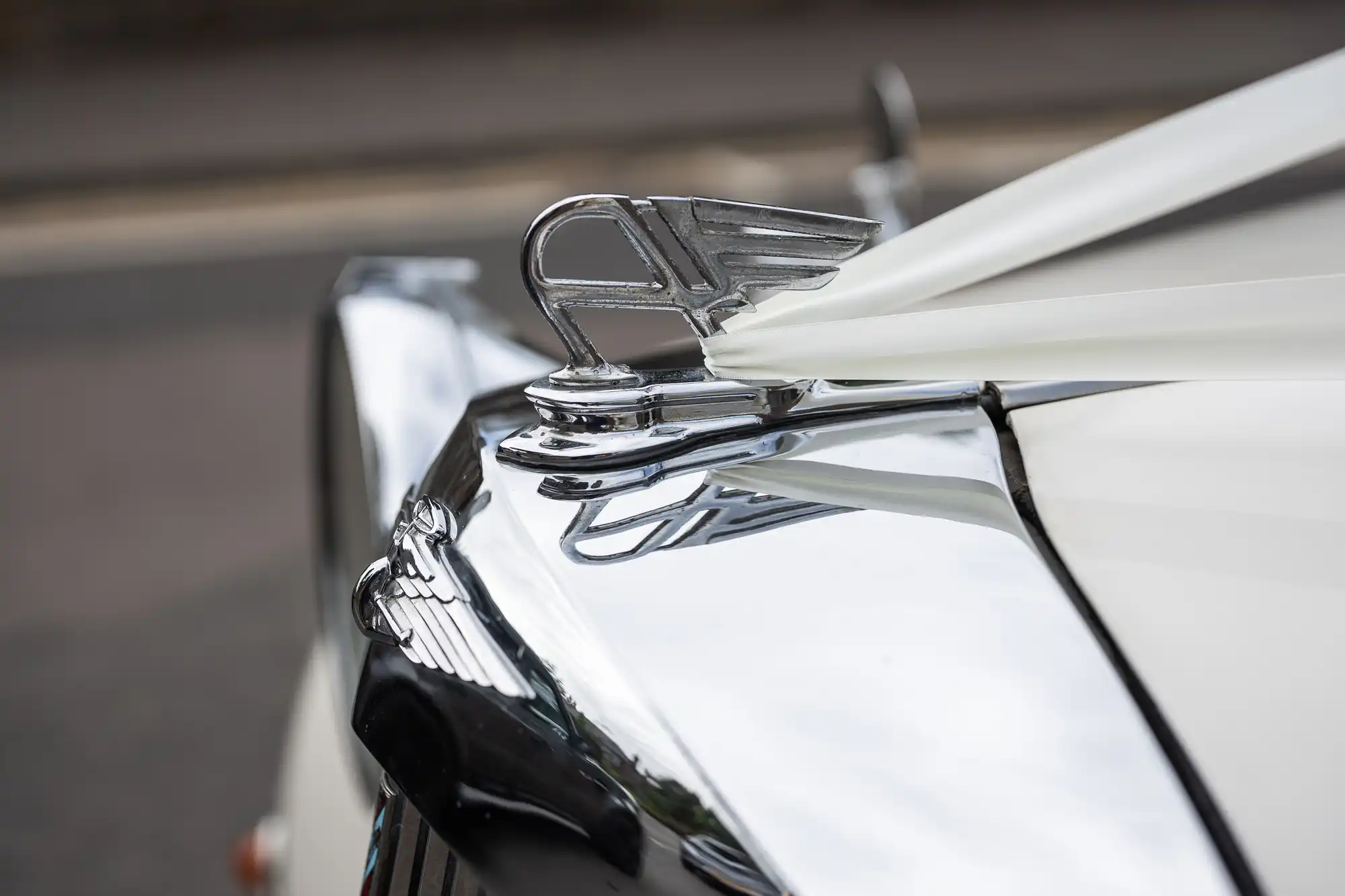 Close-up of a shiny, chrome hood ornament on a vintage car, showcasing intricate detailing against a sleek, white body.