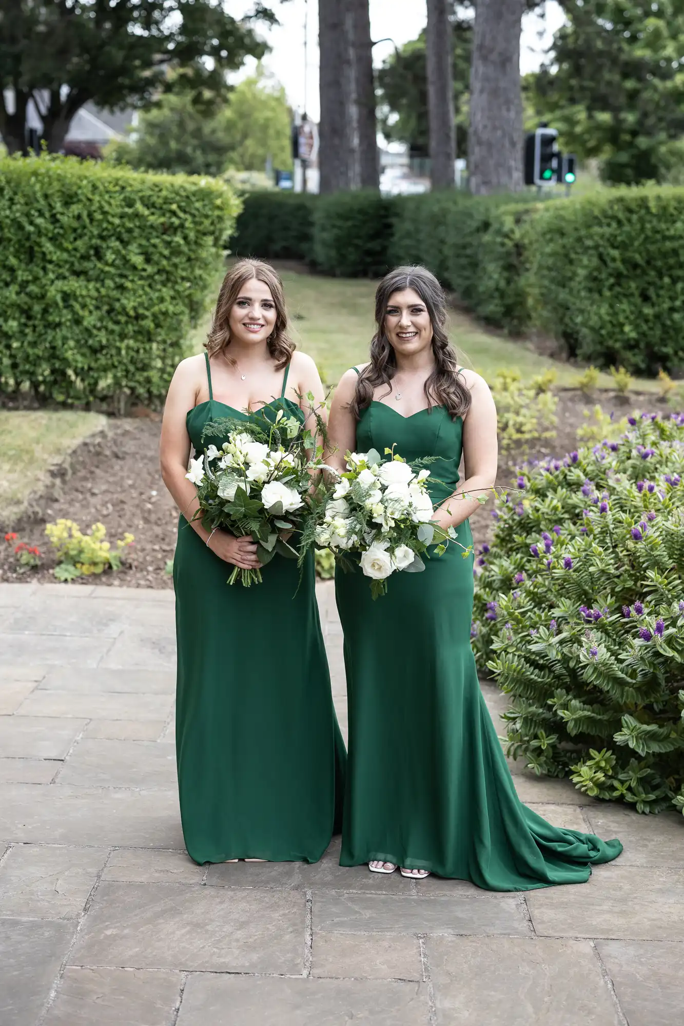 Two women in elegant green dresses holding bouquets, standing on a garden path surrounded by lush greenery.