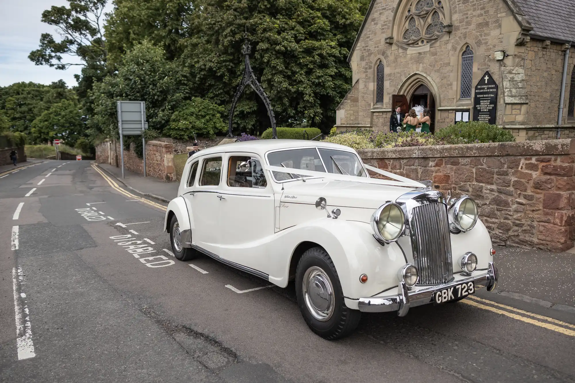 Vintage white car parked on a street next to a church, with a "Just Married" sign and decorations, and a couple visible in the church window.