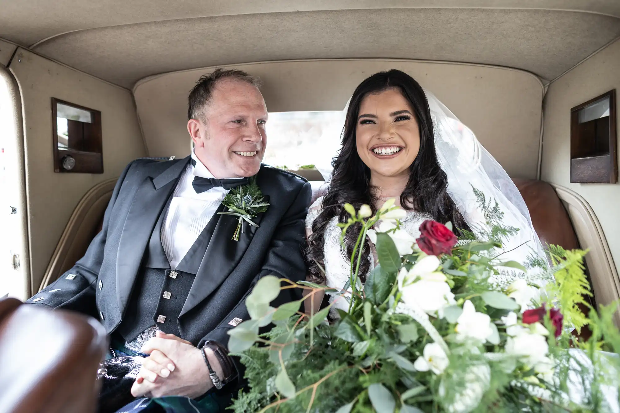 Bride and father smiling inside a vintage car, surrounded by floral decorations.