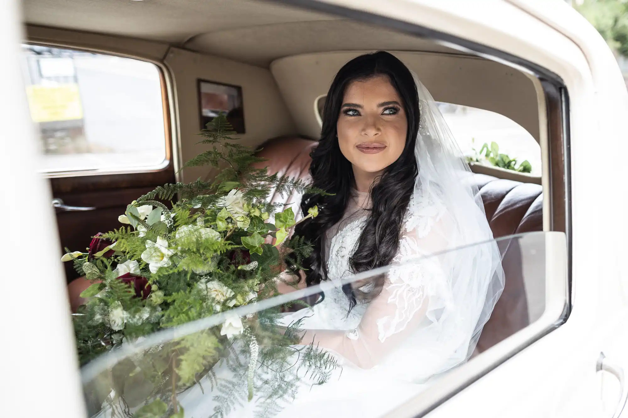 A bride holding a bouquet looks out from the window of a classic car, her expression thoughtful and serene.