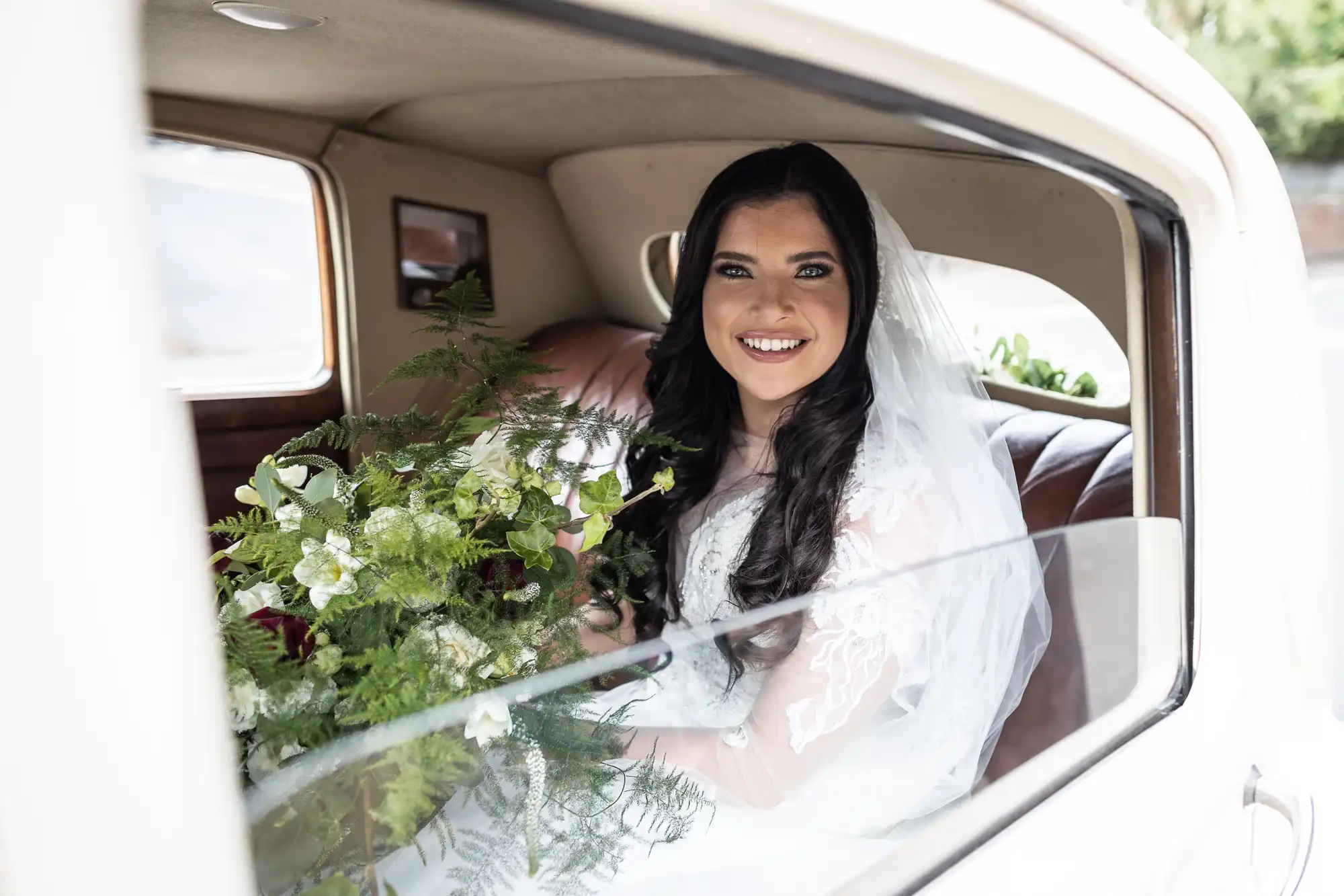 A bride smiling inside a vintage car, holding a bouquet, with natural light entering through the window.