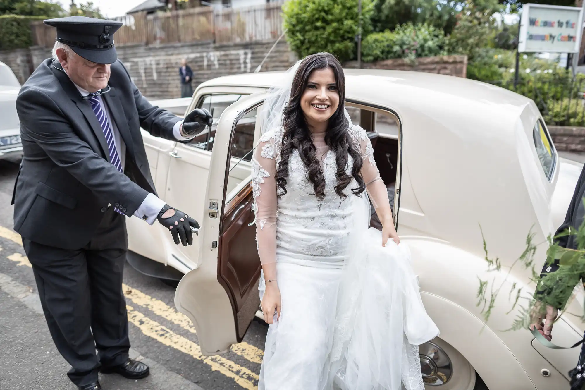 A bride in a white gown smiles as she exits a classic car with assistance from a chauffeur wearing a uniform and cap.
