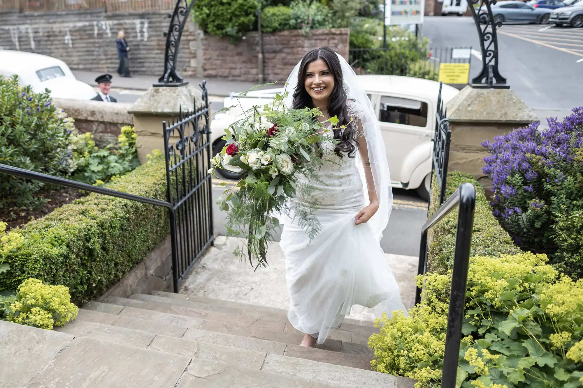 A bride smiles while walking up stone steps, holding a large bouquet, with a vintage car and chauffeur in the background.
