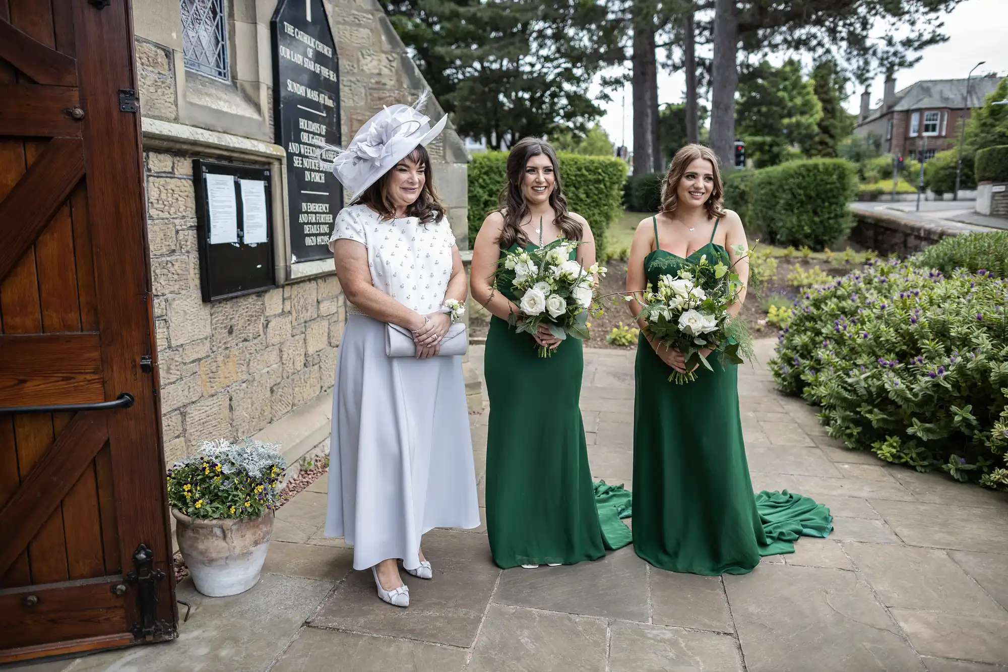 Three women in formal attire, two in green gowns and one in a white dress with a hat, holding bouquets outside a stone building.