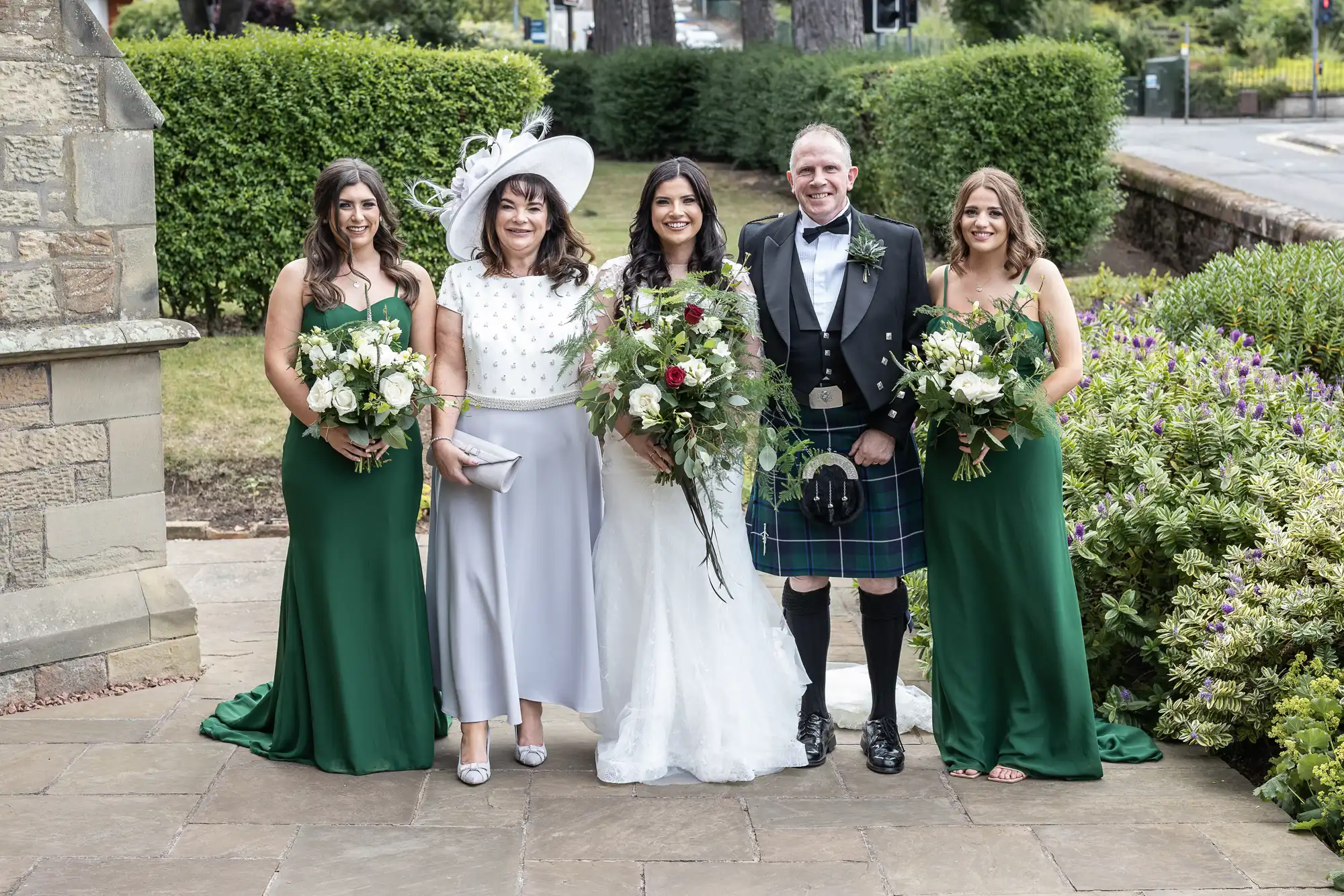 A bride and groom with three women in green dresses holding bouquets, standing by a garden. The groom wears a kilt and the bride is in a white gown.